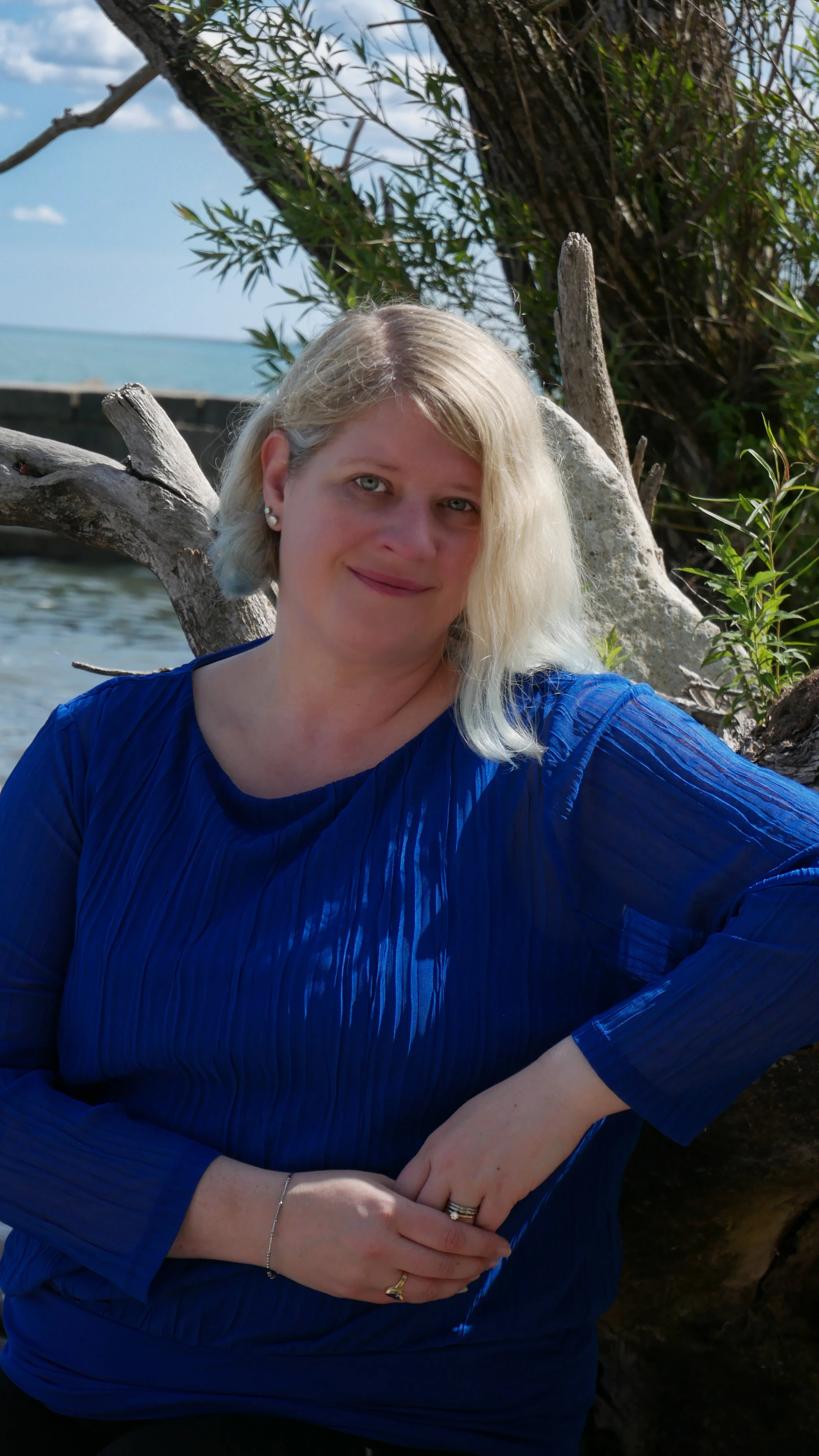 Portrait of Ashley Whitt smiling outdoors beside Lake Michigan, with trees and blue sky in the background. The photo was taken near Wind Point Lighthouse in Racine, Wisconsin. She is wearing a royal blue blouse.