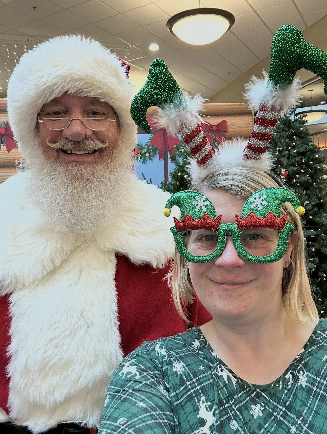 Ashley Whitt smiles for a selfie with Santa Claus while wearing festive Christmas elf glasses and a headband featuring upside-down elf legs. Decorated Christmas trees and holiday décor create a festive indoor setting.
