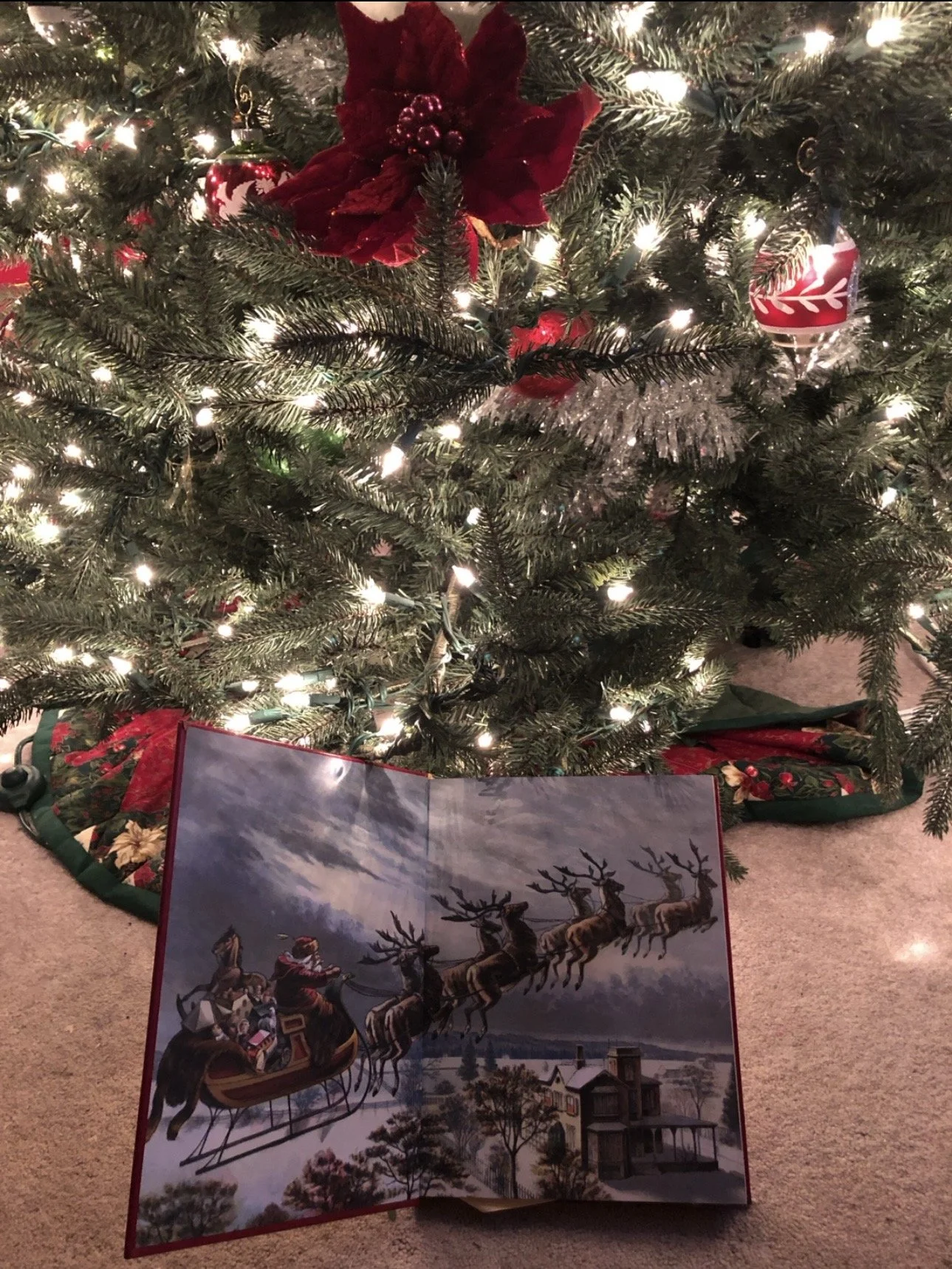 A decorated Christmas tree with white lights, red ornaments, and a large red poinsettia. Resting beneath the tree is a Christmas-themed book featuring an illustration of Santa Claus riding in a sleigh pulled by flying reindeer over a snowy landscape.