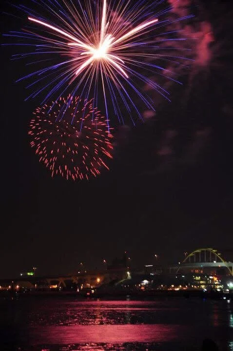 Photograph by Ashley Whitt of fireworks bursting above Milwaukee, Wisconsin, with colorful reflections shimmering across Lake Michigan at night.