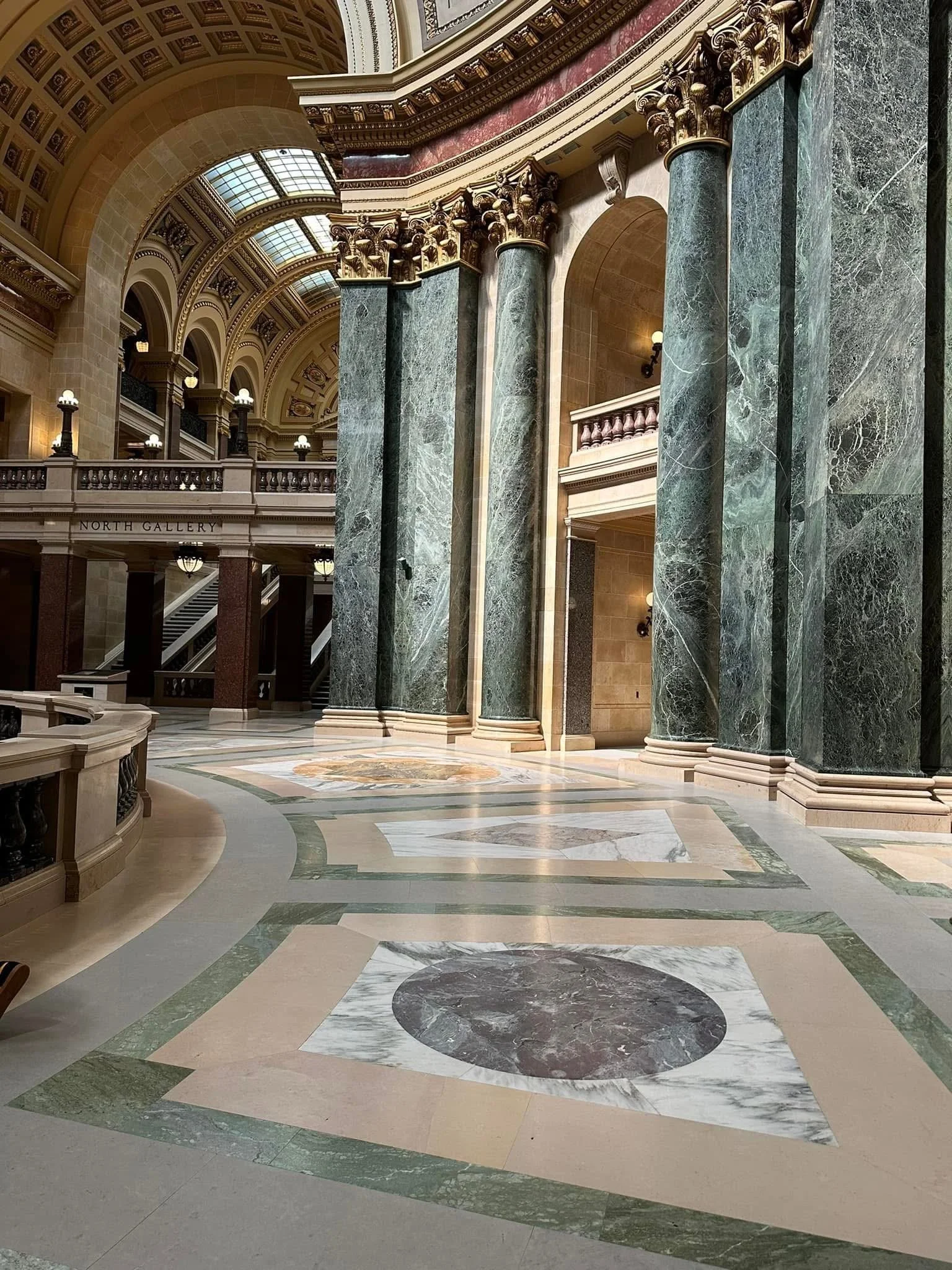 Photograph by Ashley Whitt of the interior of the Wisconsin State Capitol in Madison, Wisconsin, featuring towering marble columns, ornate arches, decorative ceilings with gold accents, and intricate marble floor patterns.