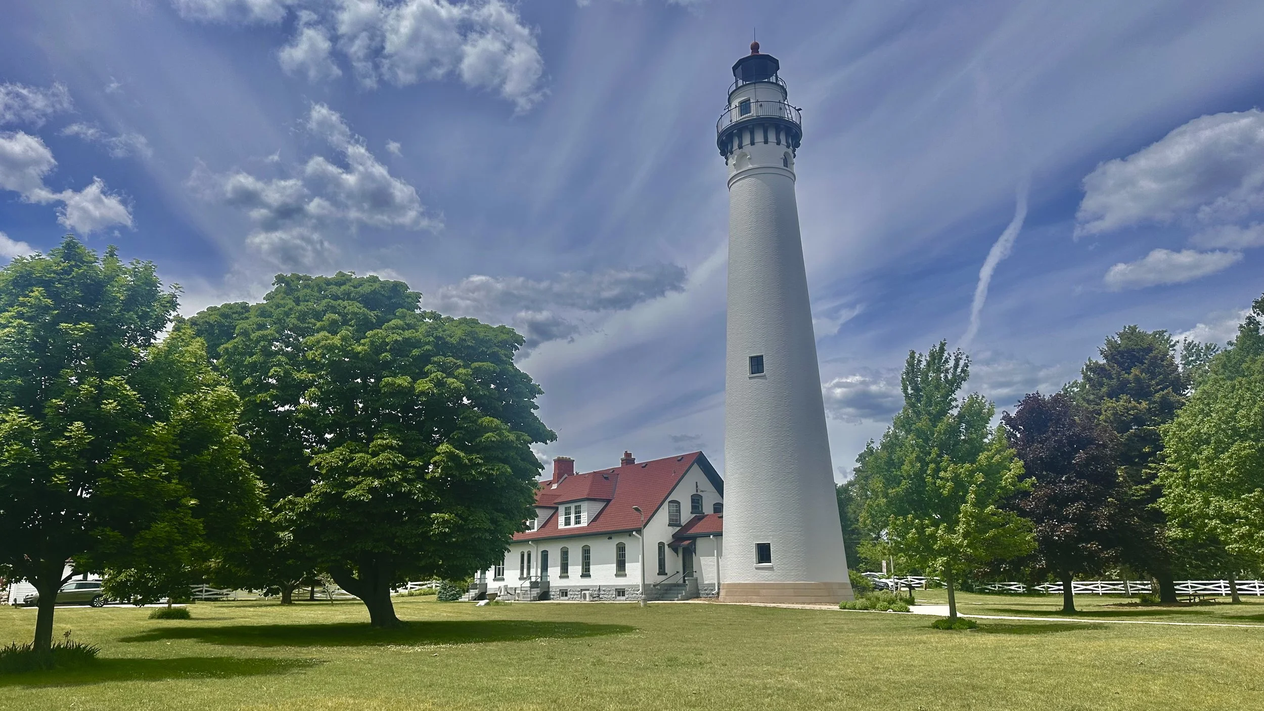 Photograph of Wind Point Lighthouse in Racine, Wisconsin, surrounded by green trees beneath a bright blue sky with scattered clouds on a sunny day.