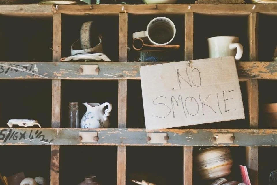 Photograph by Ashley Whitt of a rustic wooden shelf filled with vintage cups, dishes, and pottery at Hippie Tom's Serendipity Farm in Waterford, Wisconsin, featuring a handmade sign reading "NO SMOKIE."