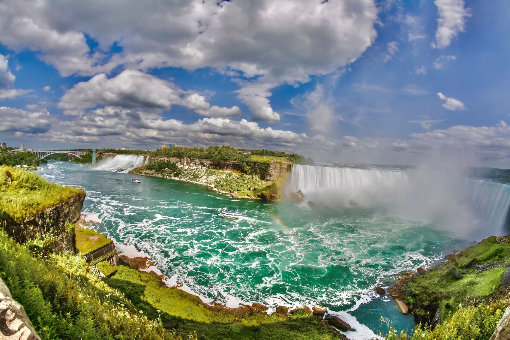 Photograph by Ashley Whitt capturing both the American Falls and Horseshoe Falls at Niagara Falls, with turquoise water, drifting mist, a rainbow, and dramatic clouds overhead.