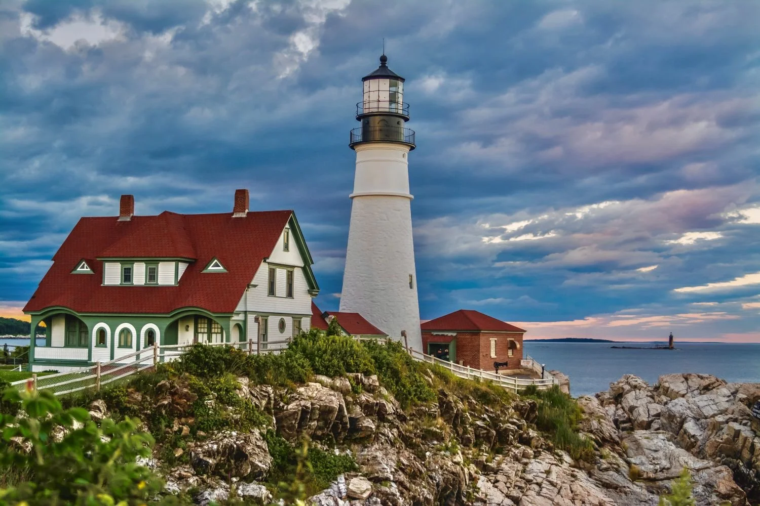 Photograph by Ashley Whitt of Portland Head Light in Cape Elizabeth, Maine, overlooking the rocky Atlantic coastline beneath dramatic clouds with patches of blue sky.