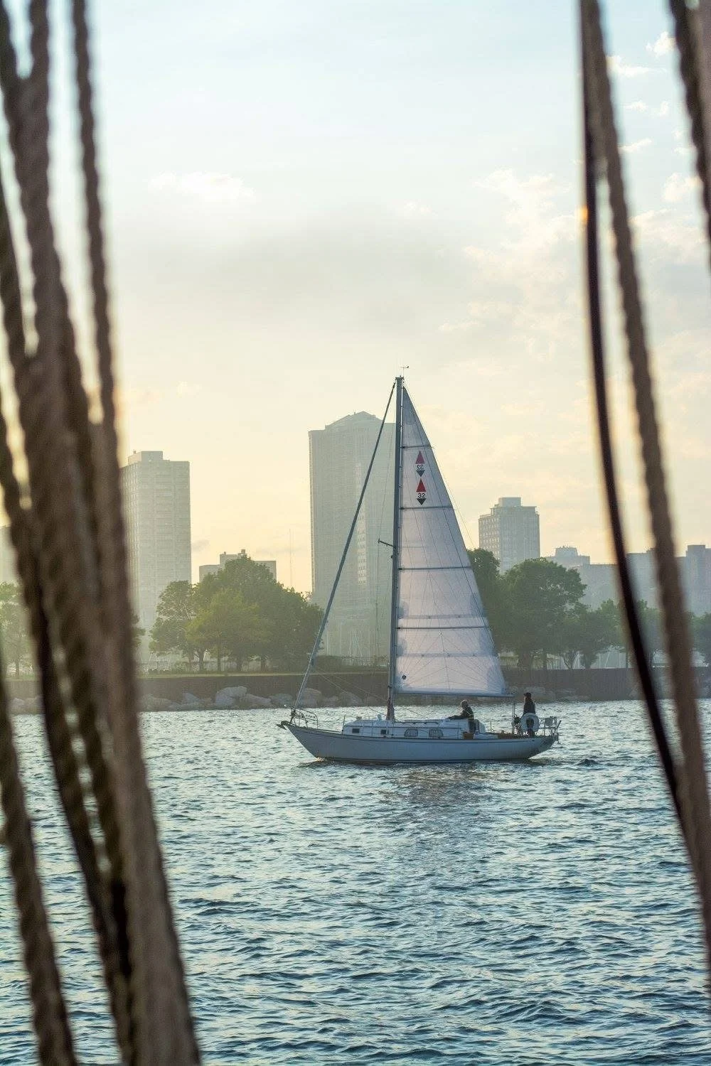 Photograph by Ashley Whitt of a sailboat gliding across Lake Michigan near the Wisconsin shoreline during sunset, viewed through the rigging of a historic tall ship.