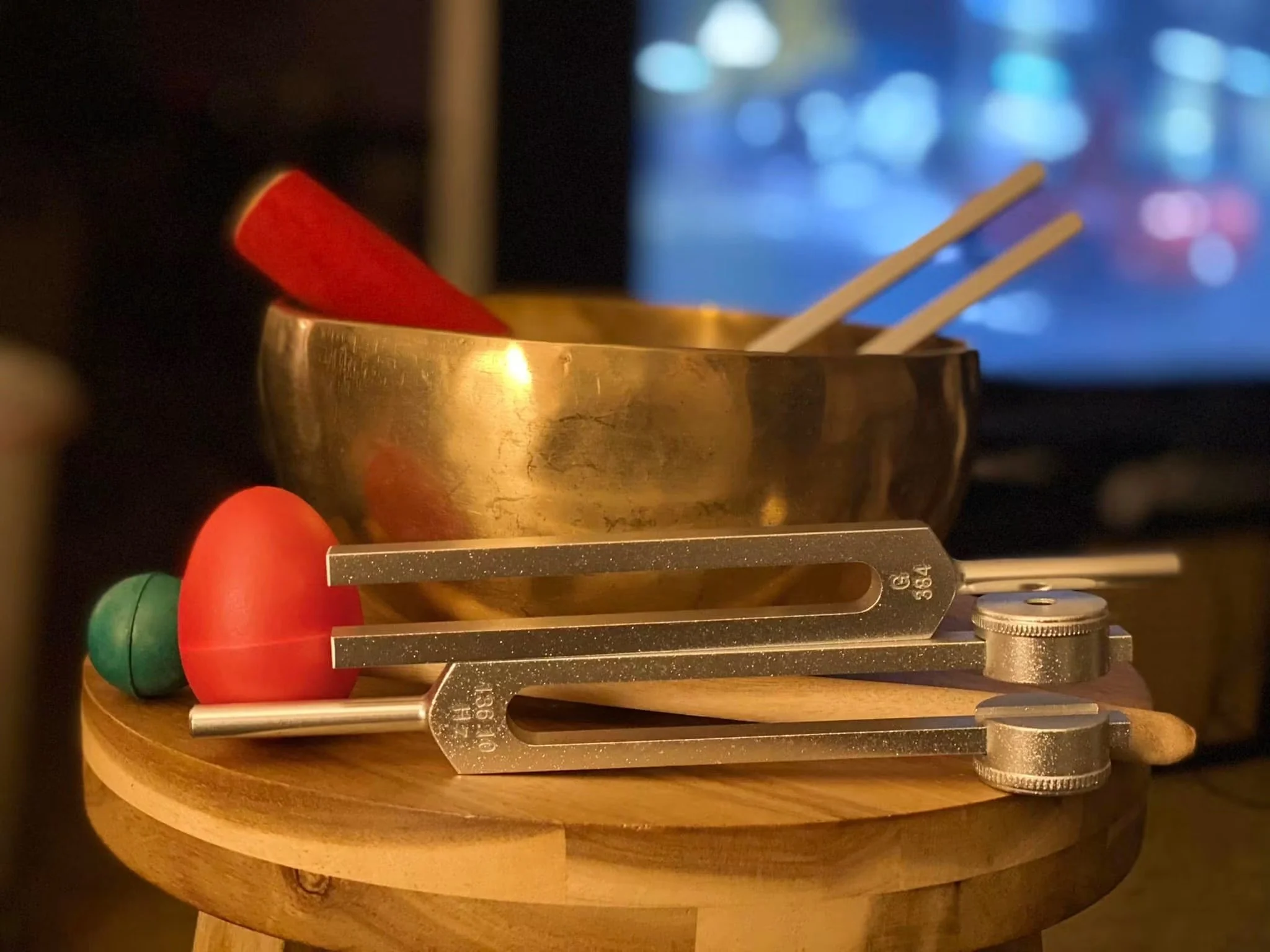Close-up photograph of sound healing tools including tuning forks, mallets, red egg shaker, and brass singing bowl arranged on a wooden surface with a softly blurred background.