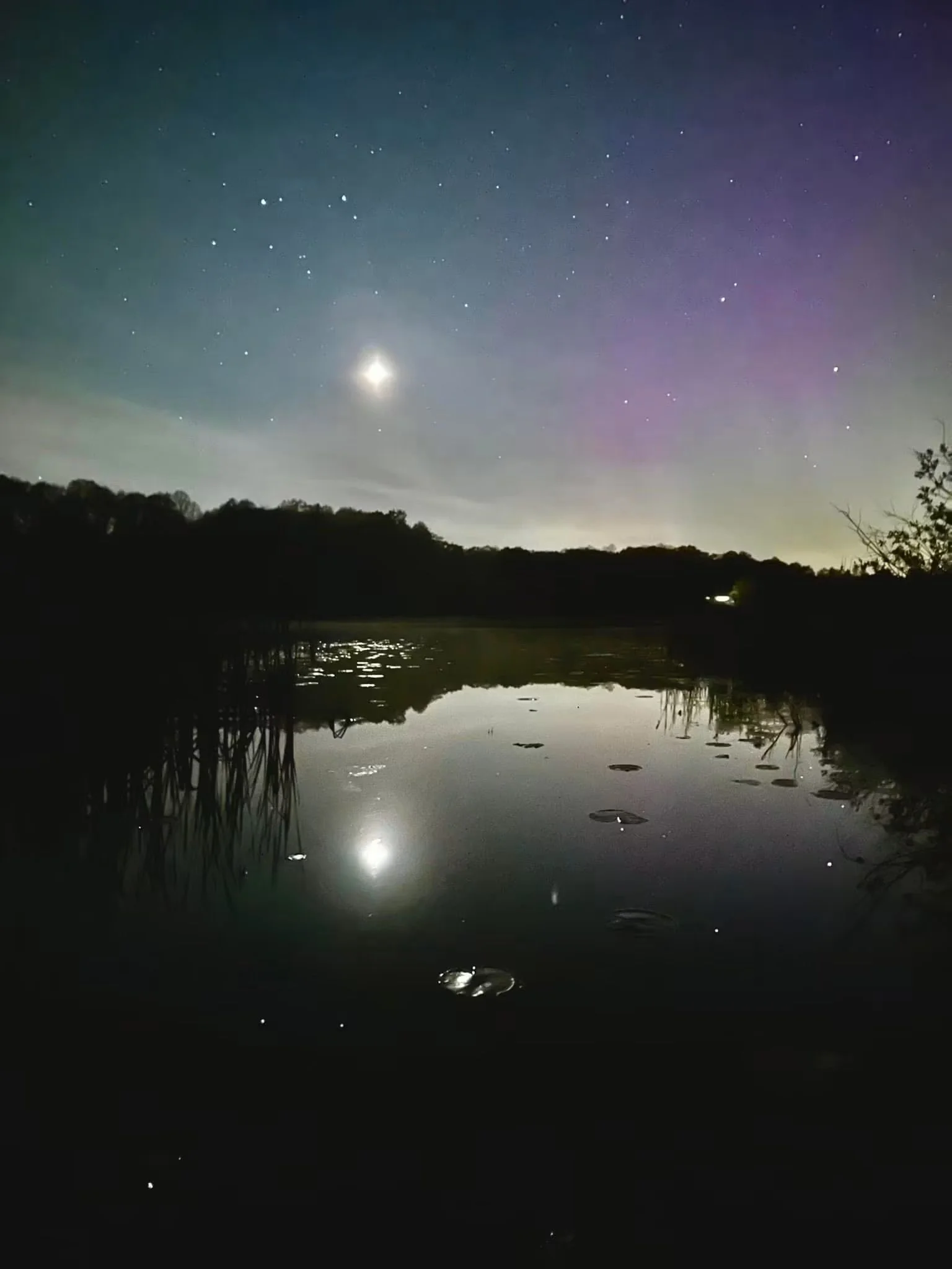 Photograph by Ashley Whitt of the northern lights glowing above calm water in Scott, Wisconsin, with reflections of the moon, stars, and colorful aurora shimmering across the still surface.