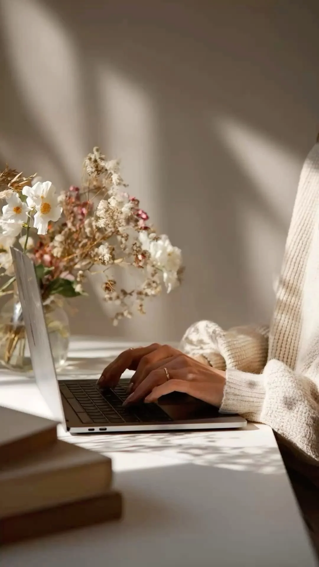 Person typing on a laptop with a bouquet of flowers and books on the desk.