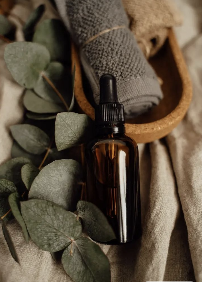A brown glass dropper bottle beside eucalyptus leaves, a rolled gray towel, and a wooden dish with a cloth inside, all arranged on beige fabric.