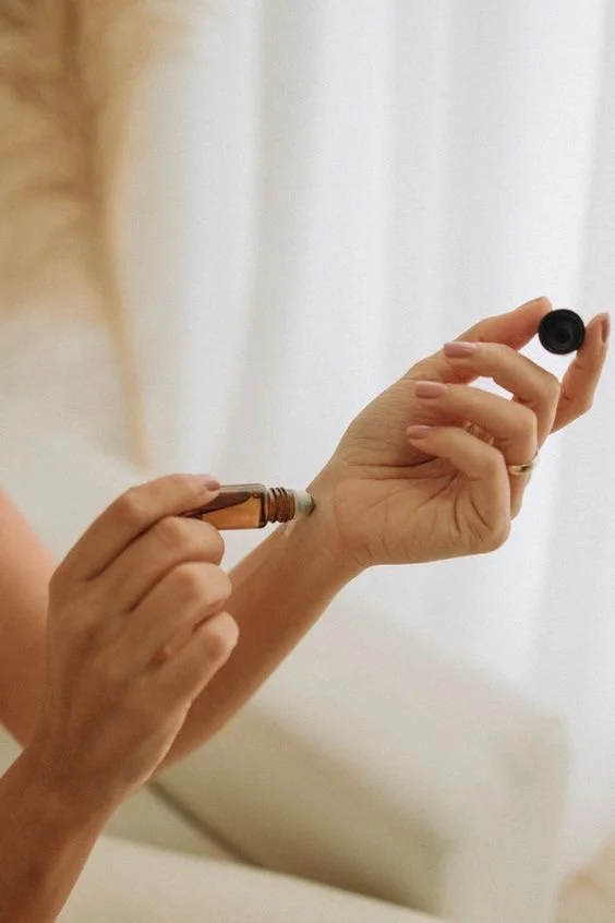 A person is holding a small bottle or tube in one hand and squeezing the contents onto their other hand, which is holding a black cap, in front of a white curtain background.