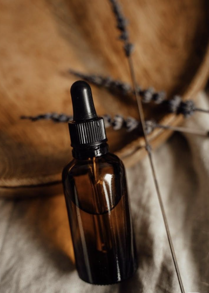 A small amber glass dropper bottle with a black screw cap and rubber dropper, placed on a textured surface with blurred natural-colored fabric and a pin cushion with pins in the background.