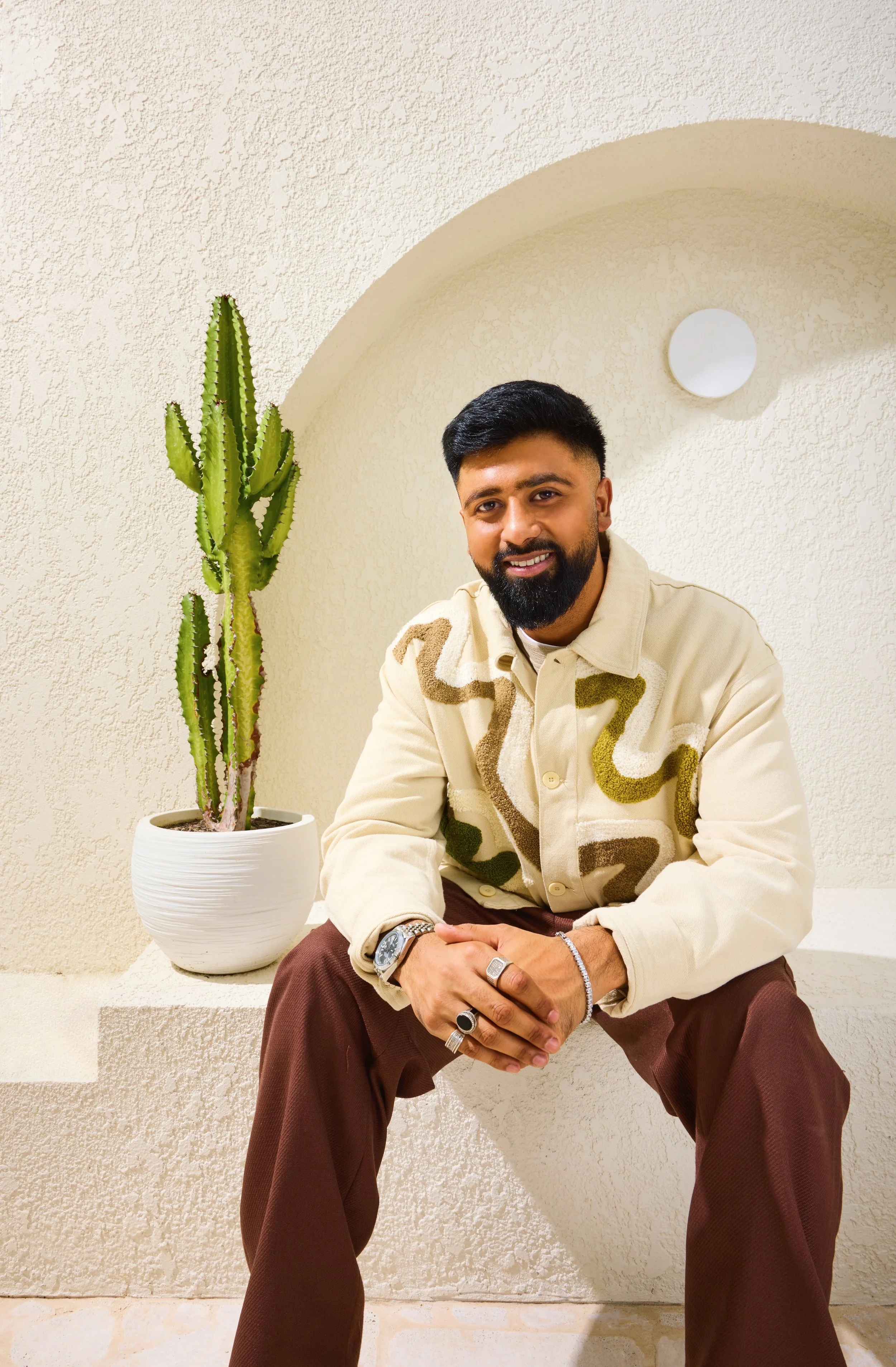 A man with black hair, a beard, and multiple rings on his fingers, wearing a beige patterned jacket and brown pants, sitting on a step next to a potted cactus plant against a textured beige wall.