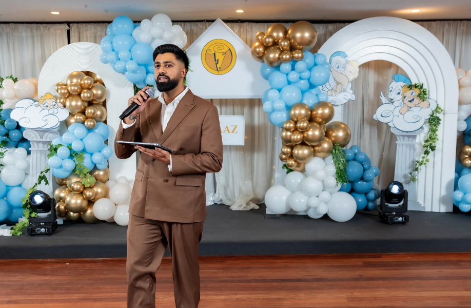 A man in a brown suit holding a microphone and a folder speaking at a decorated event stage with cartoon clouds and gold, blue, and white balloons.