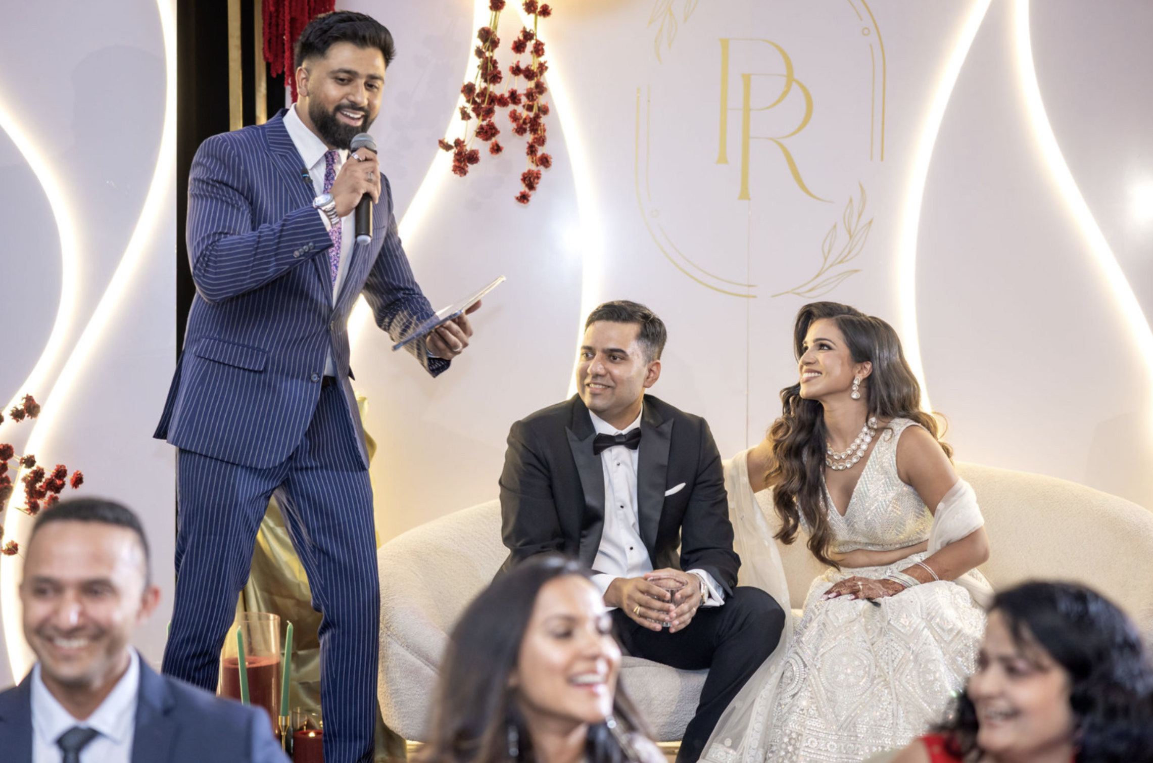 A man in a blue pinstripe suit is giving a speech while holding a microphone and a paper. Seated around him are well-dressed people, including a woman in a traditional white patterned dress with jewelry and a man in a black tuxedo with a bowtie. The setting appears to be a formal event or celebration, decorated with flowers and a white and gold backdrop.