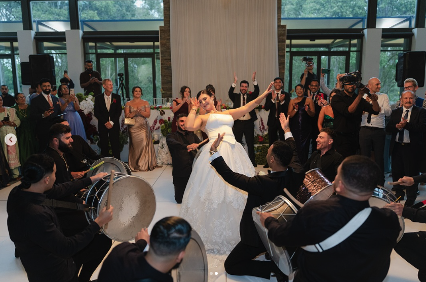 Bride dancing with live musicians playing drums at her wedding celebration, surrounded by guests clapping and watching in a modern indoor venue.