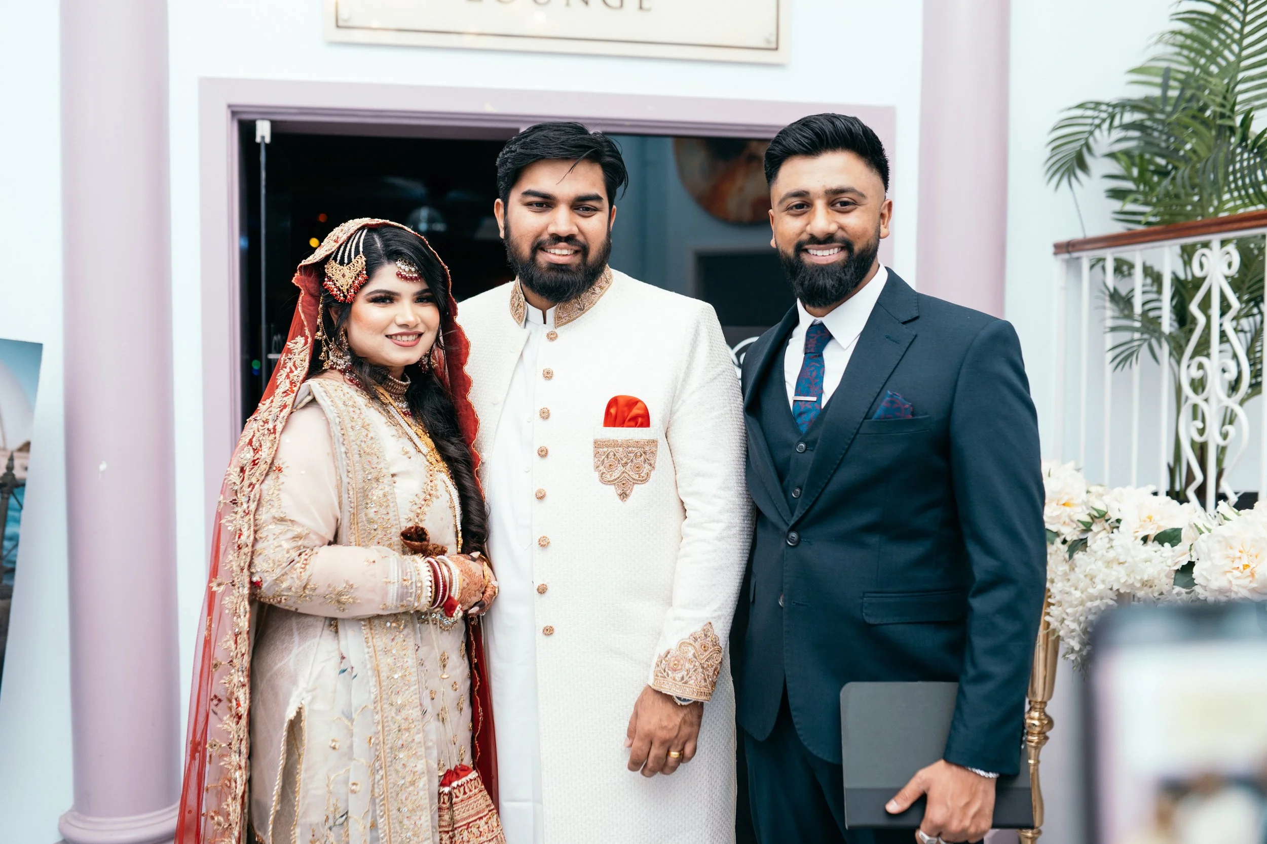 Three people dressed in formal attire, with a woman in traditional South Asian wedding clothing, a man in a beige sherwani, and another man in a dark suit, smiling together at a celebration, with flowers and greenery in the background.