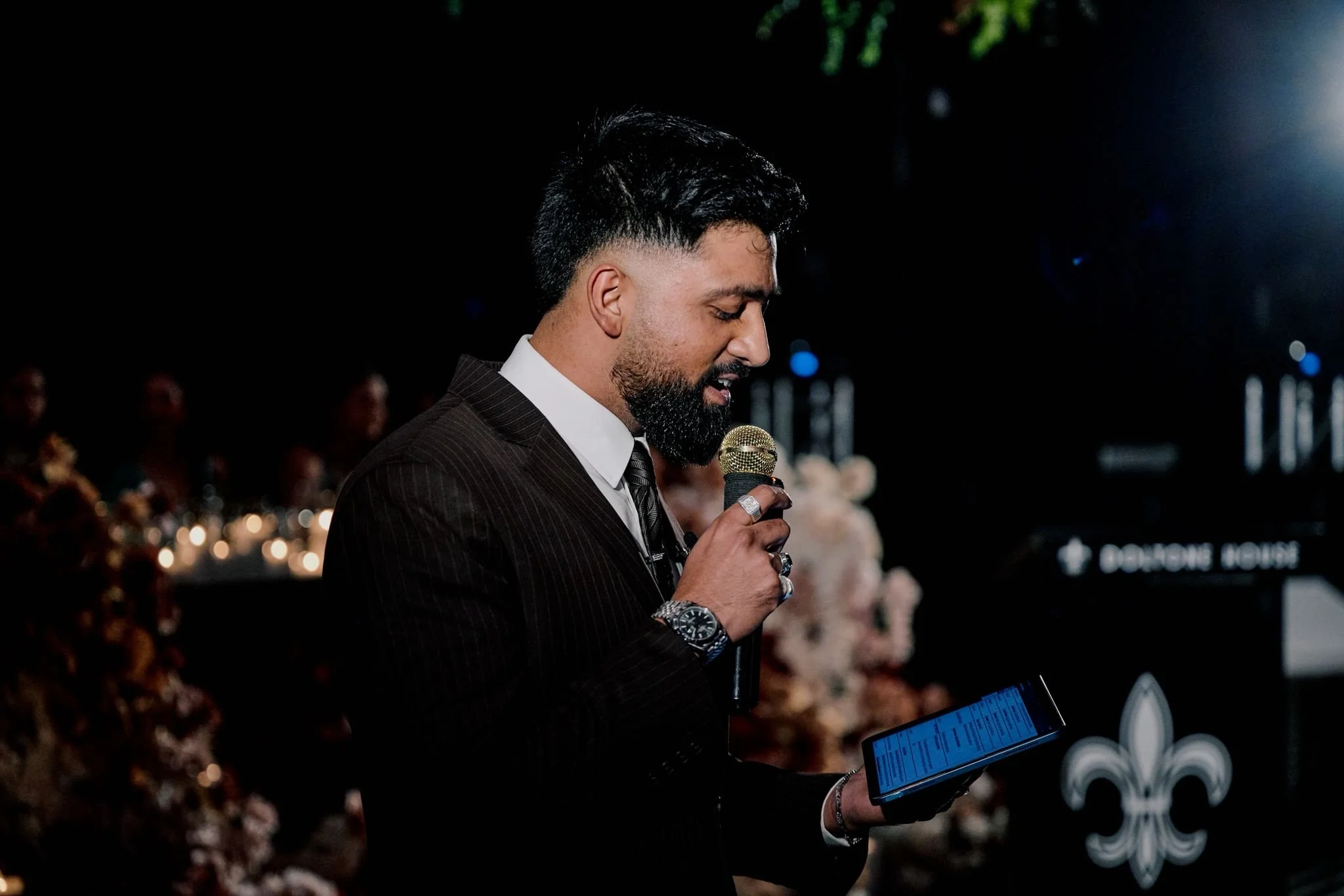 A man in a dark pinstripe suit and white shirt is holding a microphone and a tablet, speaking at an indoor event with floral decorations and floral-themed backdrop.