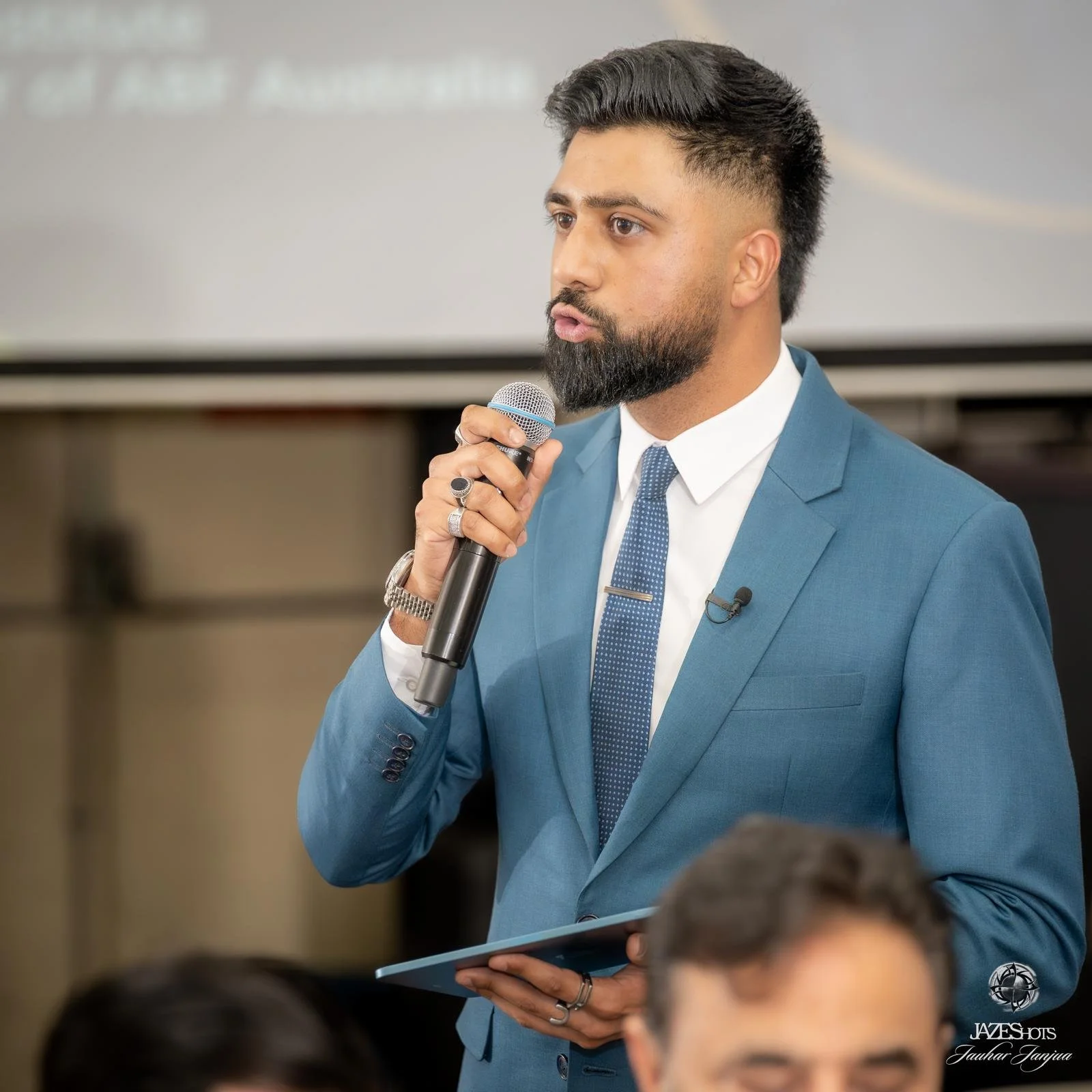 A man in a blue suit holding a microphone and a folder appears to be speaking at a conference or meeting.