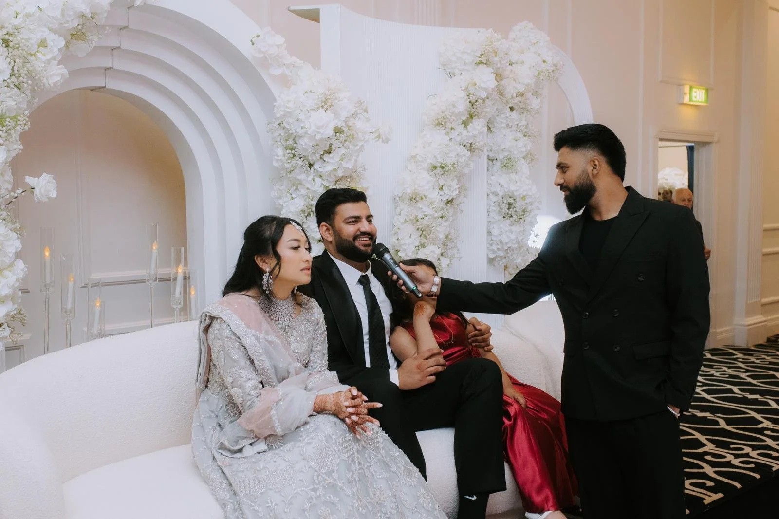 Group of four people at a wedding, one woman in traditional attire with henna on her hands, a man in a suit and tie, a woman in a red dress, and a man in a black suit, seated on a white couch with floral decorations behind them, as a man with a microphone interviews the seated individuals.