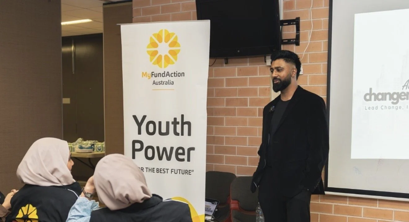 A man standing in front of a presentation screen and a banner that reads 'Youth Power' with a logo for MyFundAction Australia, in a room with brick walls. Two women in headscarves are seated and listening.