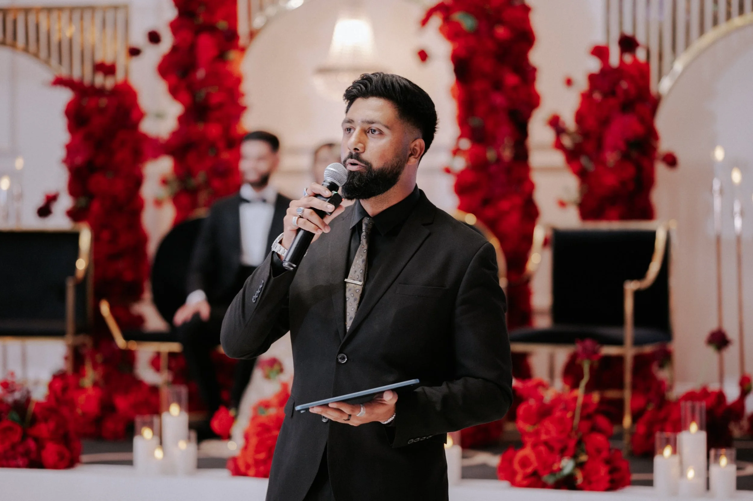 A man with dark hair and beard wearing a black suit and tie, speaking into a microphone at an event with a decorated background of red flowers and candles.