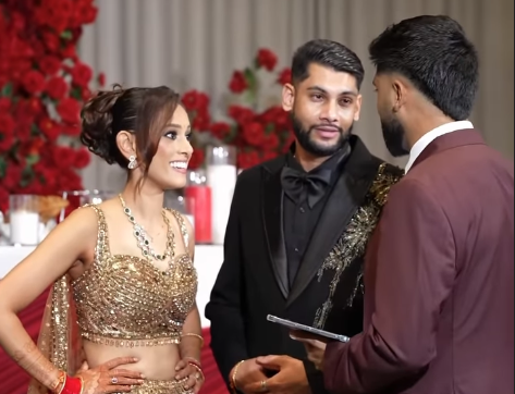 Three people dressed in formal attire at a wedding reception, with a woman in a gold dress and jewelry and two men in suits, one holding a tablet, engaged in conversation in front of a decorated backdrop with red flowers.