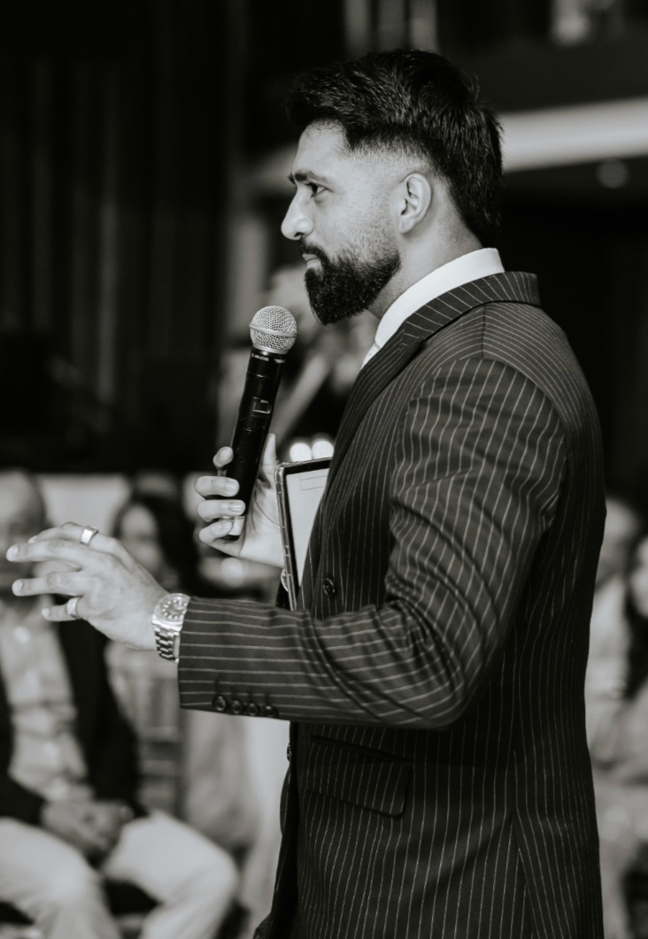 A man with a beard and styled hair wearing a pinstripe suit is speaking into a microphone during a presentation or speech in a professional setting.