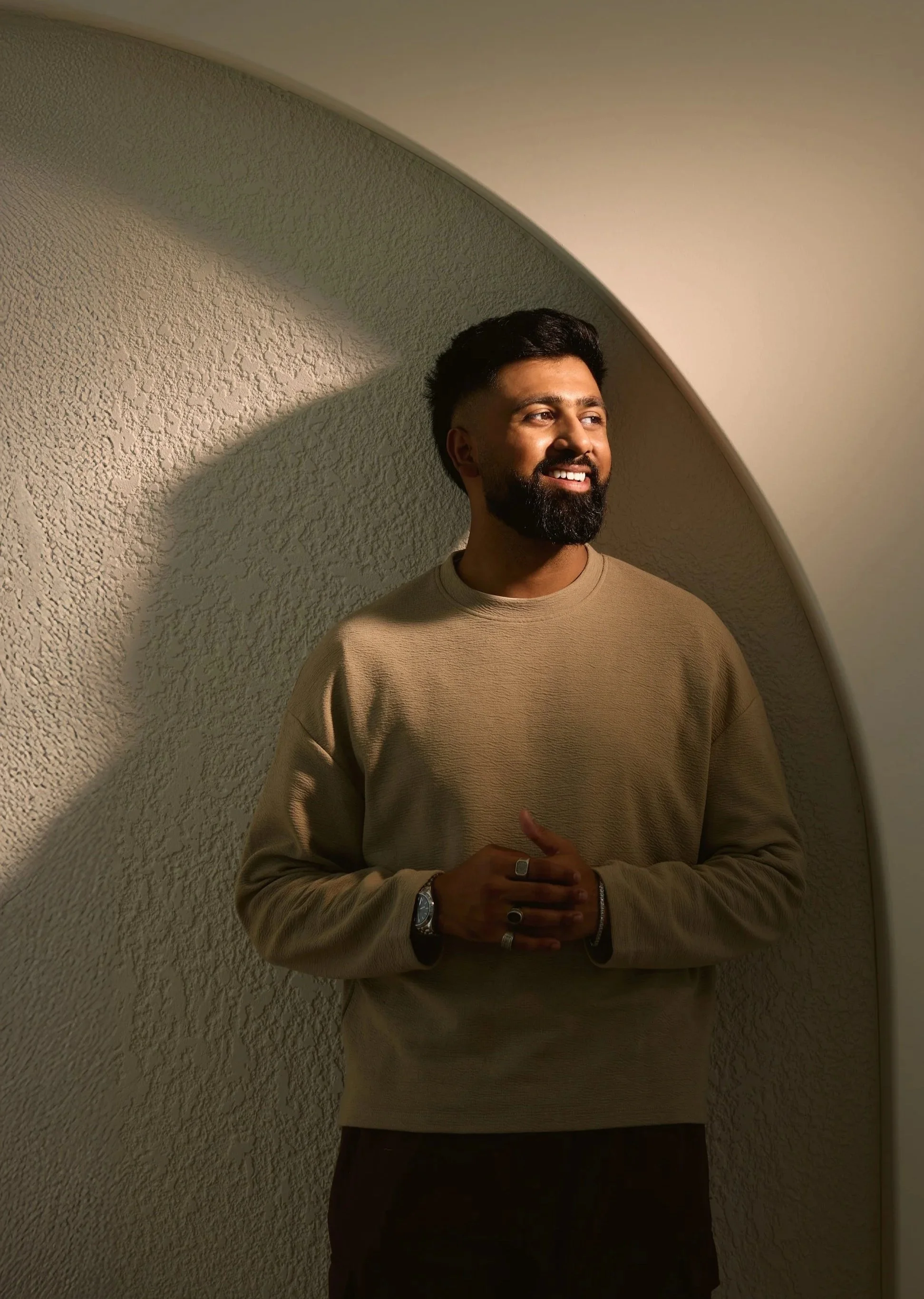 A man with dark hair and a beard standing against a textured, curved wall, smiling and looking to his right. He is wearing a beige sweater, a watch, and rings, with his hands clasped in front of him.