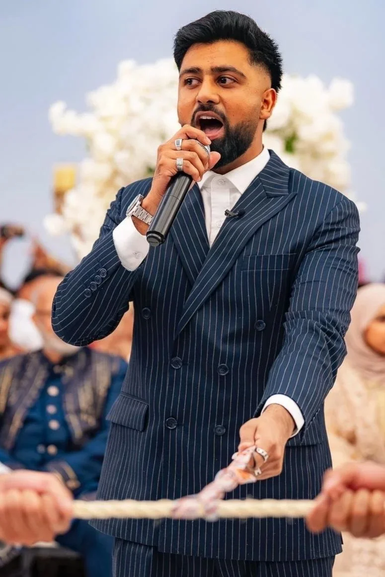 A man in a navy blue pinstripe suit speaking into a microphone during a formal event. People in the background are seated and wearing traditional attire.