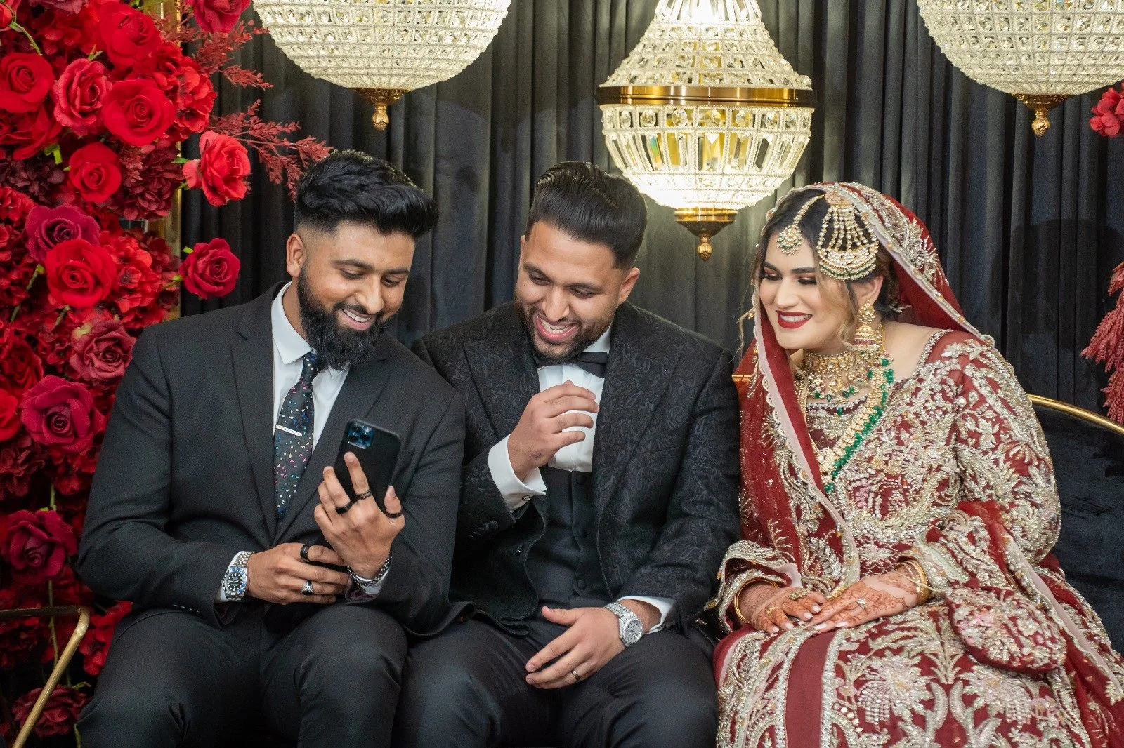 Three people sitting on a sofa, dressed in formal and traditional Indian wedding attire, looking at a smartphone and smiling.