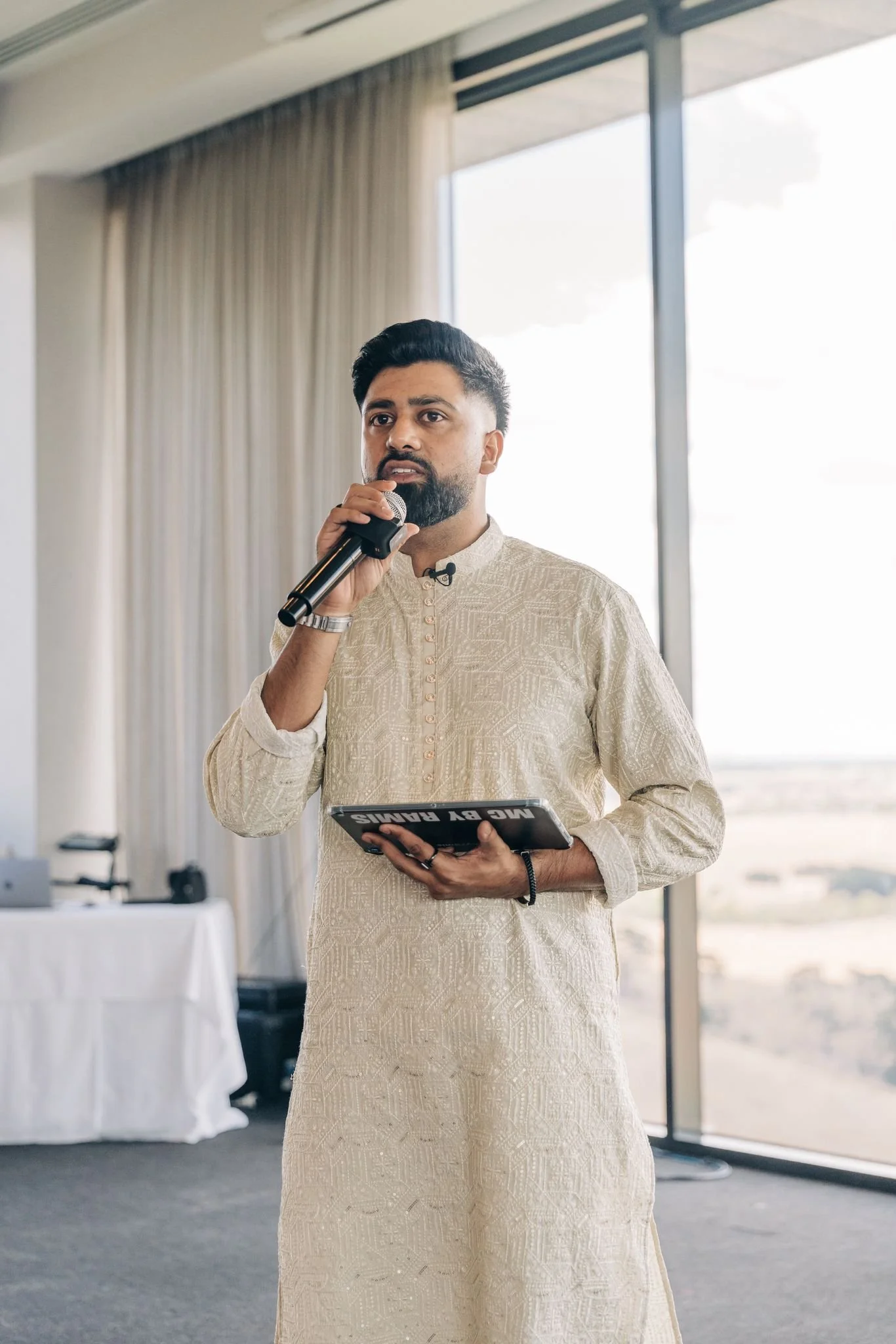 A man dressed in traditional cream-colored Indian attire, holding a microphone and a tablet, speaking at an indoor event near large windows with curtains.