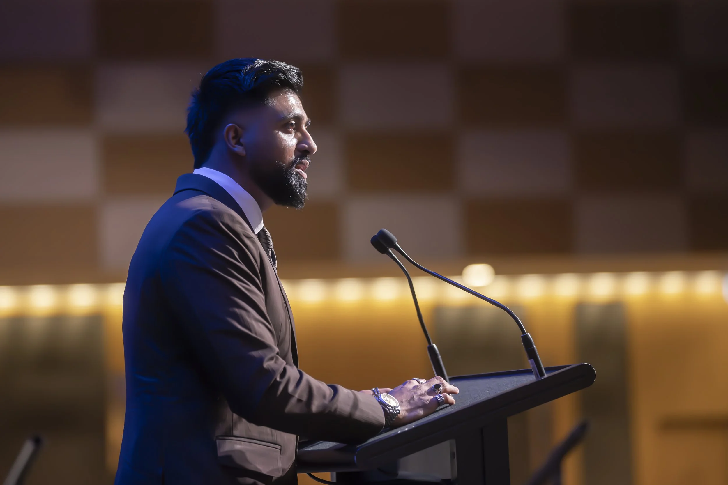 A man with dark hair and a beard stands at a podium, wearing a suit and tie, speaking into a microphone in a modern auditorium with a warm, softly lit background.