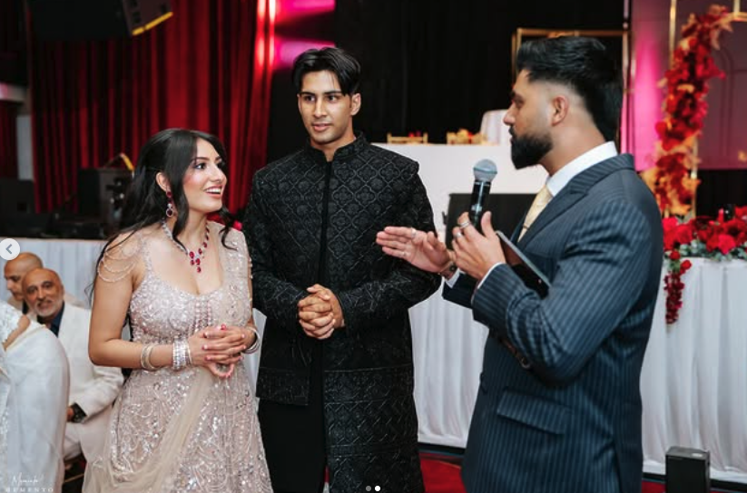 A couple in traditional Indian attire standing and listening to a man in a suit holding a microphone during a celebration or wedding reception.