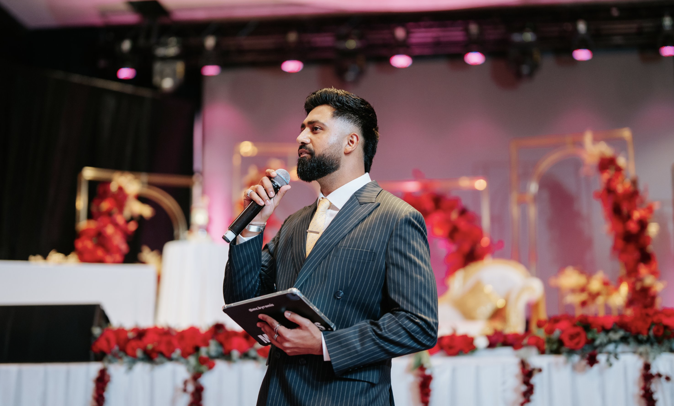 A man with a beard in a pinstripe suit holding a microphone and reading from a notepad at an event with red and gold floral decorations and pink overhead lighting.