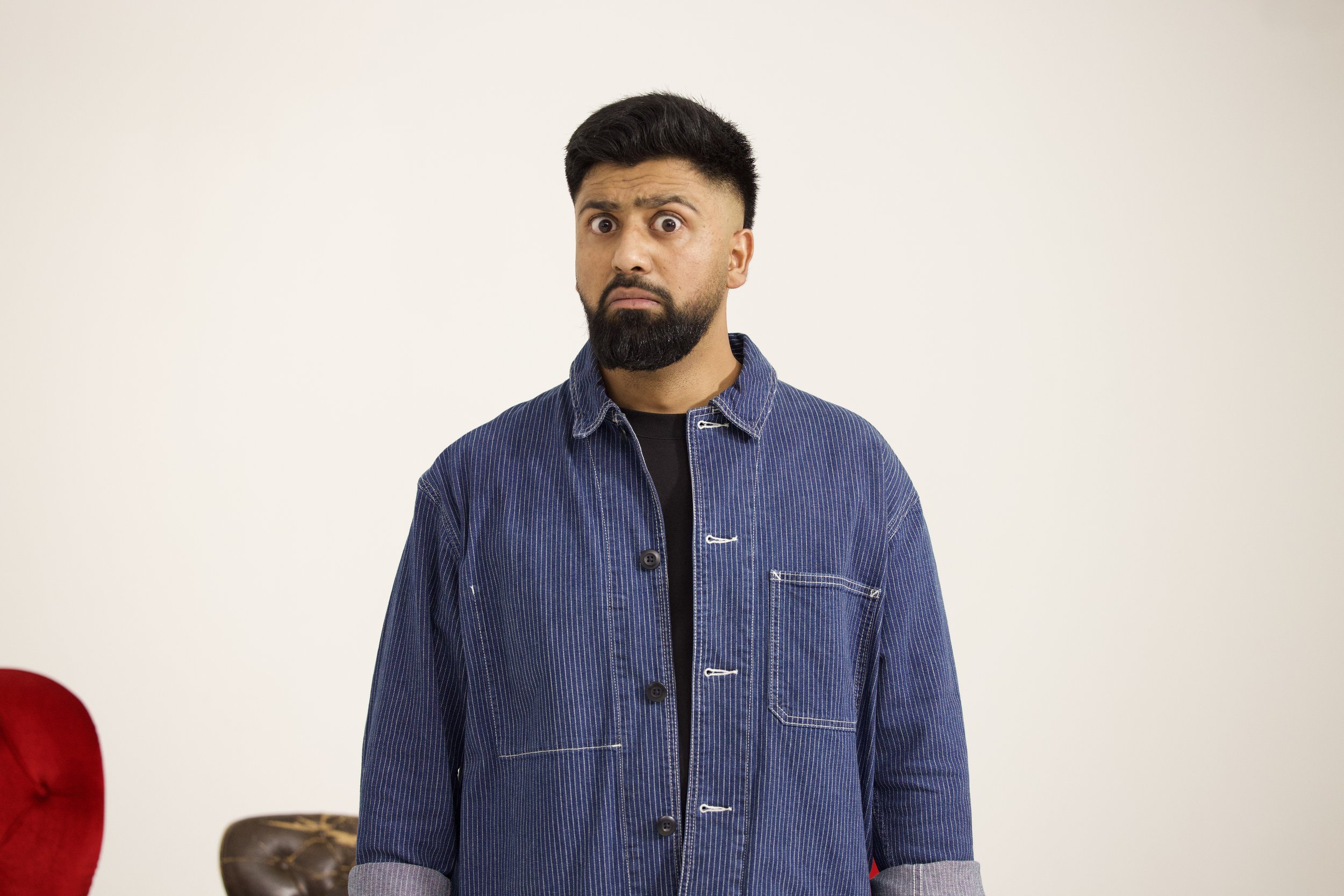 A man with dark hair, a beard, and mustache, wearing a blue denim shirt with rolled-up sleeves, stands against a plain light background with a surprised or confused expression on his face.