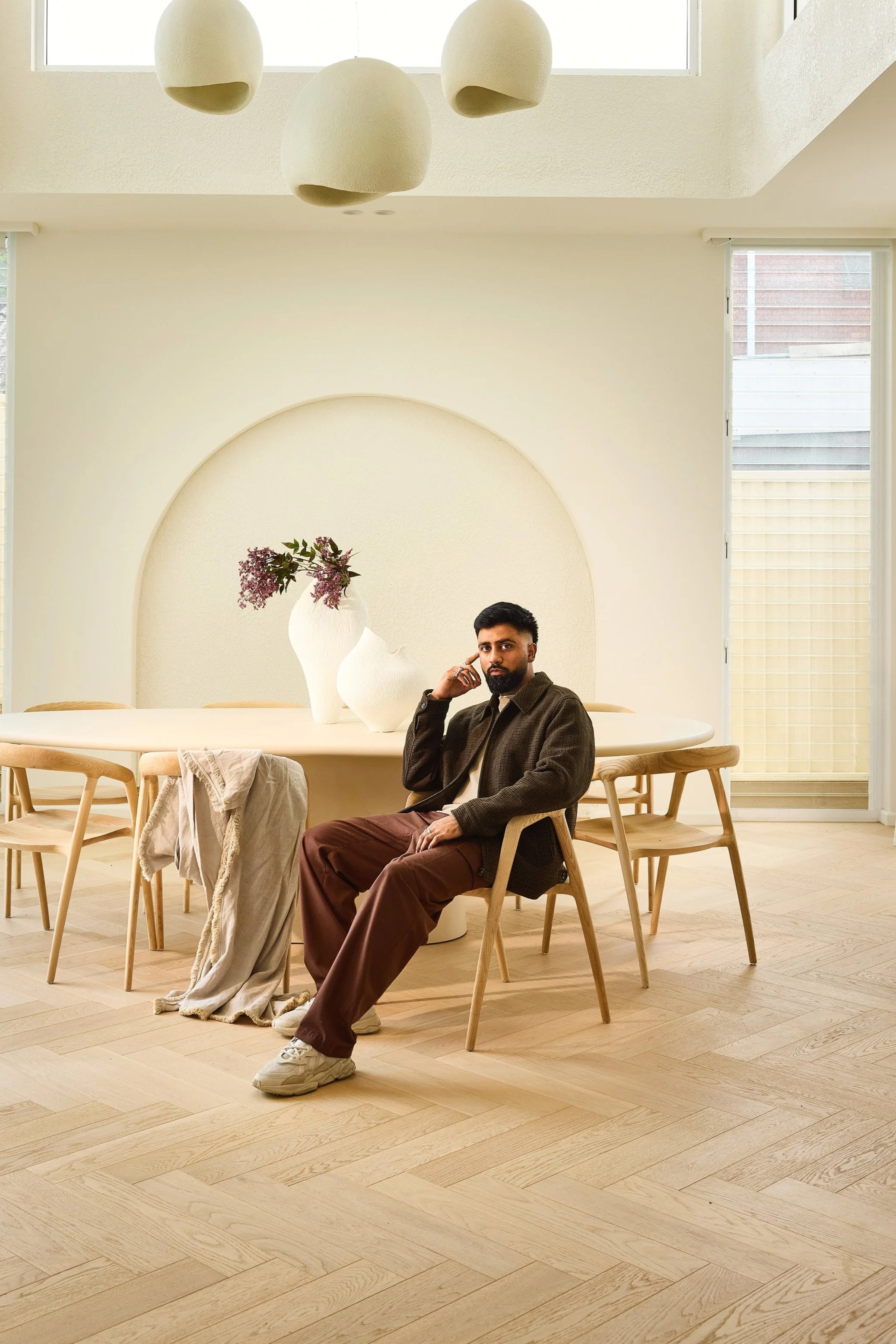 A man sitting at a round wooden table in a bright, minimalist dining room with natural light, dressed in brown pants, sneakers, and a dark jacket, with beige chairs and white vases with flowers on the table.