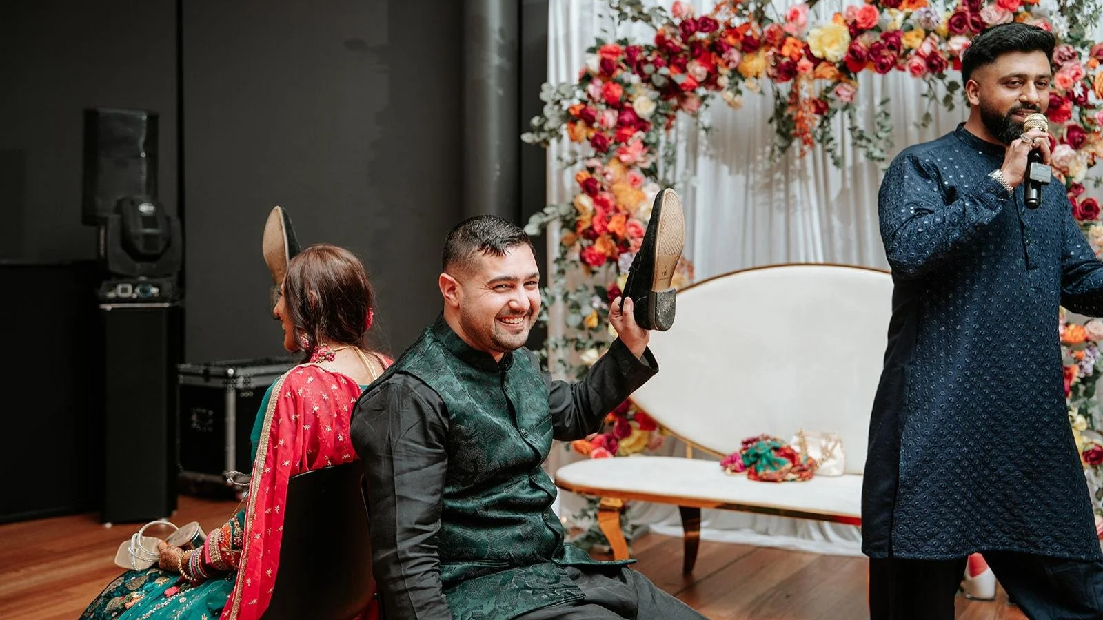 People celebrating at a wedding or event with a floral backdrop, including a man smiling and holding a shoe, a woman dressed in traditional attire, and a man speaking into a microphone.