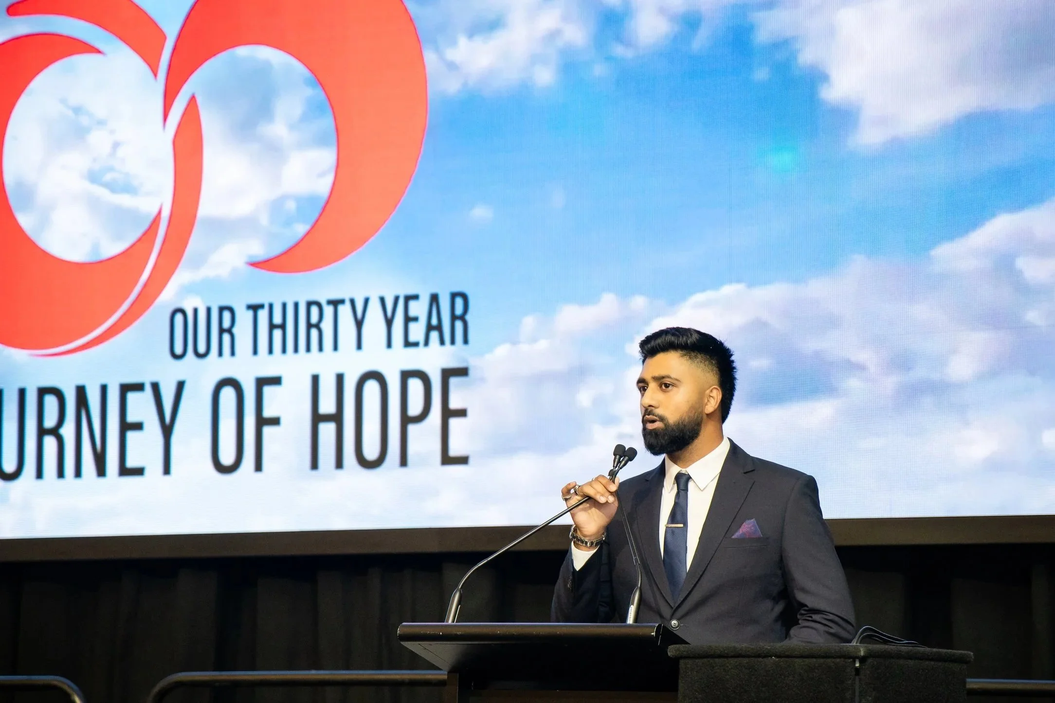 A man in a dark suit and tie speaking at a podium, holding a microphone, with a large screen behind him displaying a red logo and the text 'Our Thirty Year Journey of Hope' against a blue sky with clouds.
