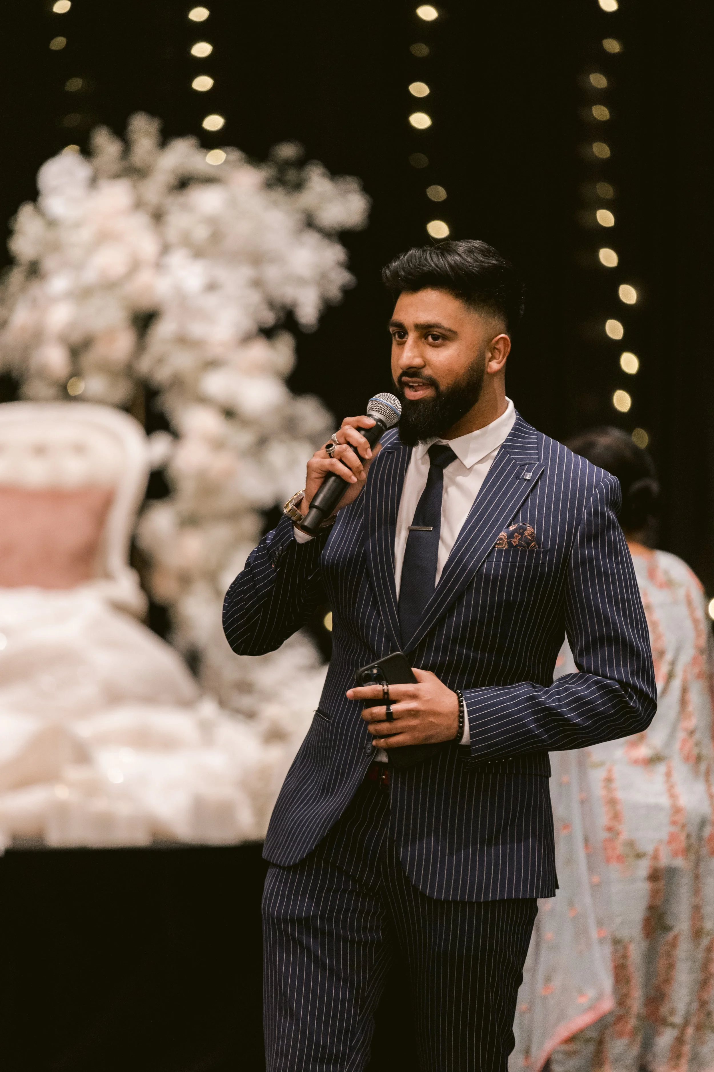 A man in a blue pinstripe suit holding a microphone and a phone, speaking at an event decorated with blurred white flowers and fairy lights in the background.