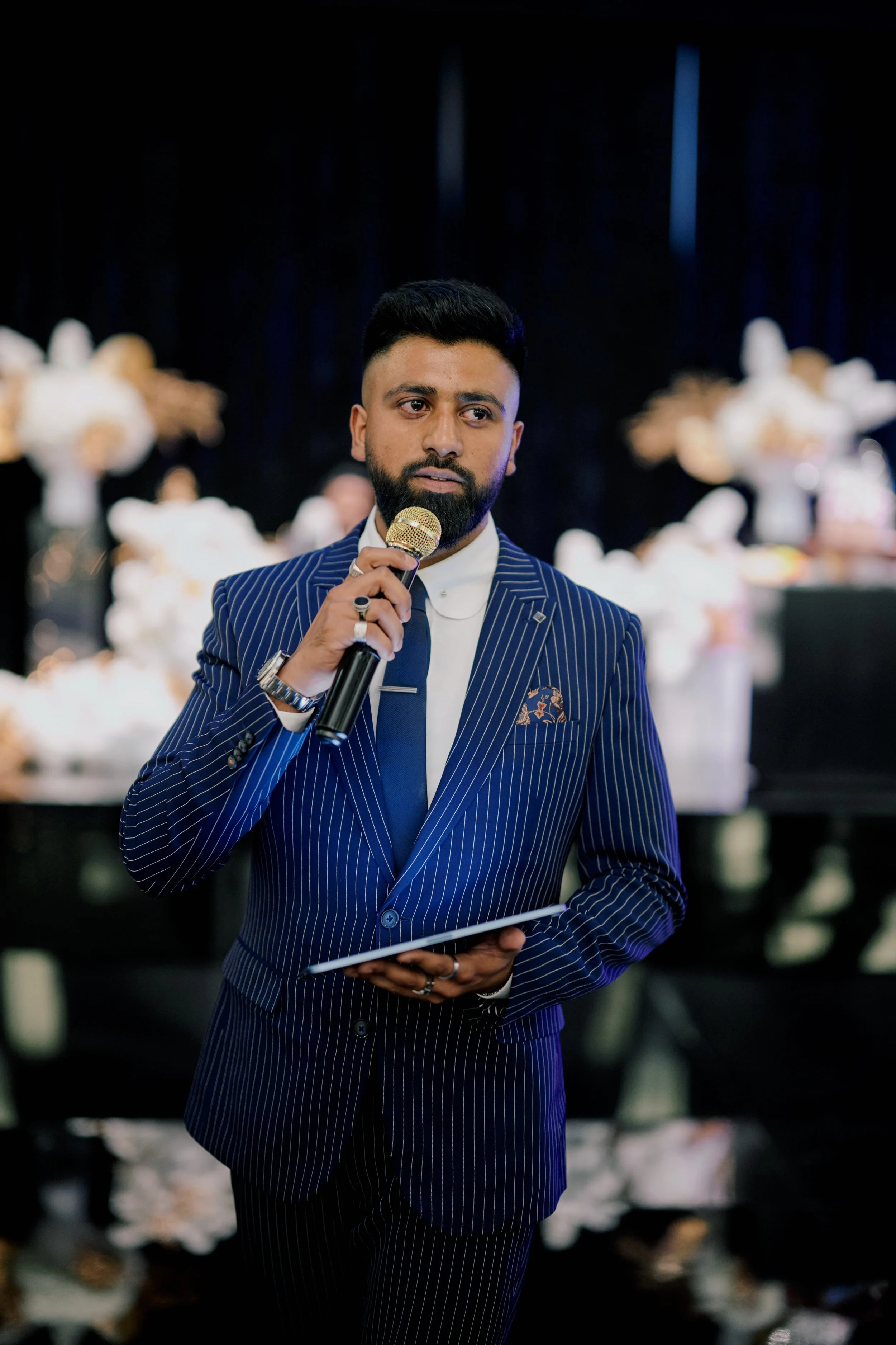 Man with black hair and beard wearing a pinstripe suit holding a microphone at an event