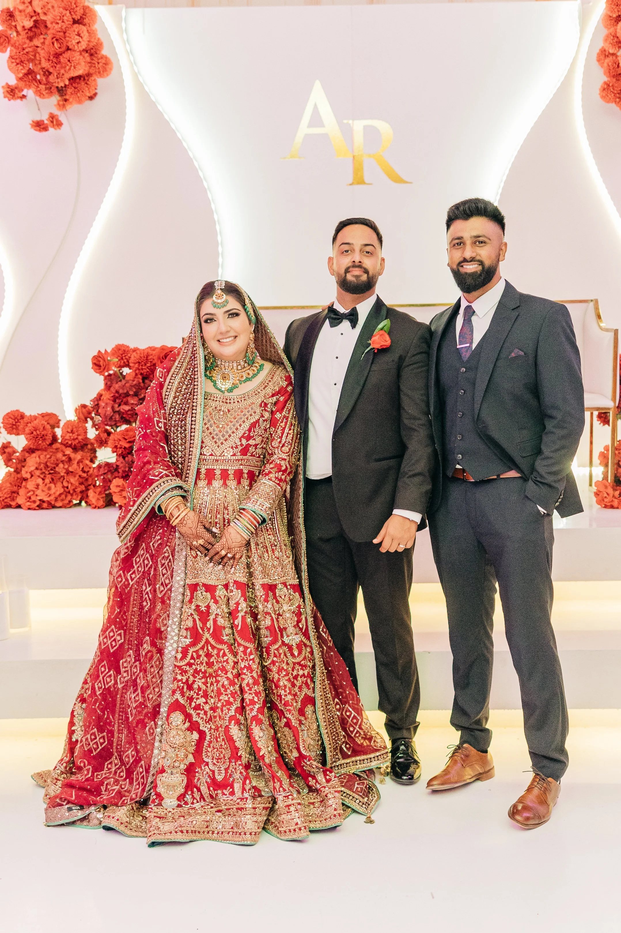 A bride in traditional red and gold Indian wedding attire and jewelry standing with two men in black suits at a wedding reception with a decorated backdrop featuring red flowers and initials 'AR'