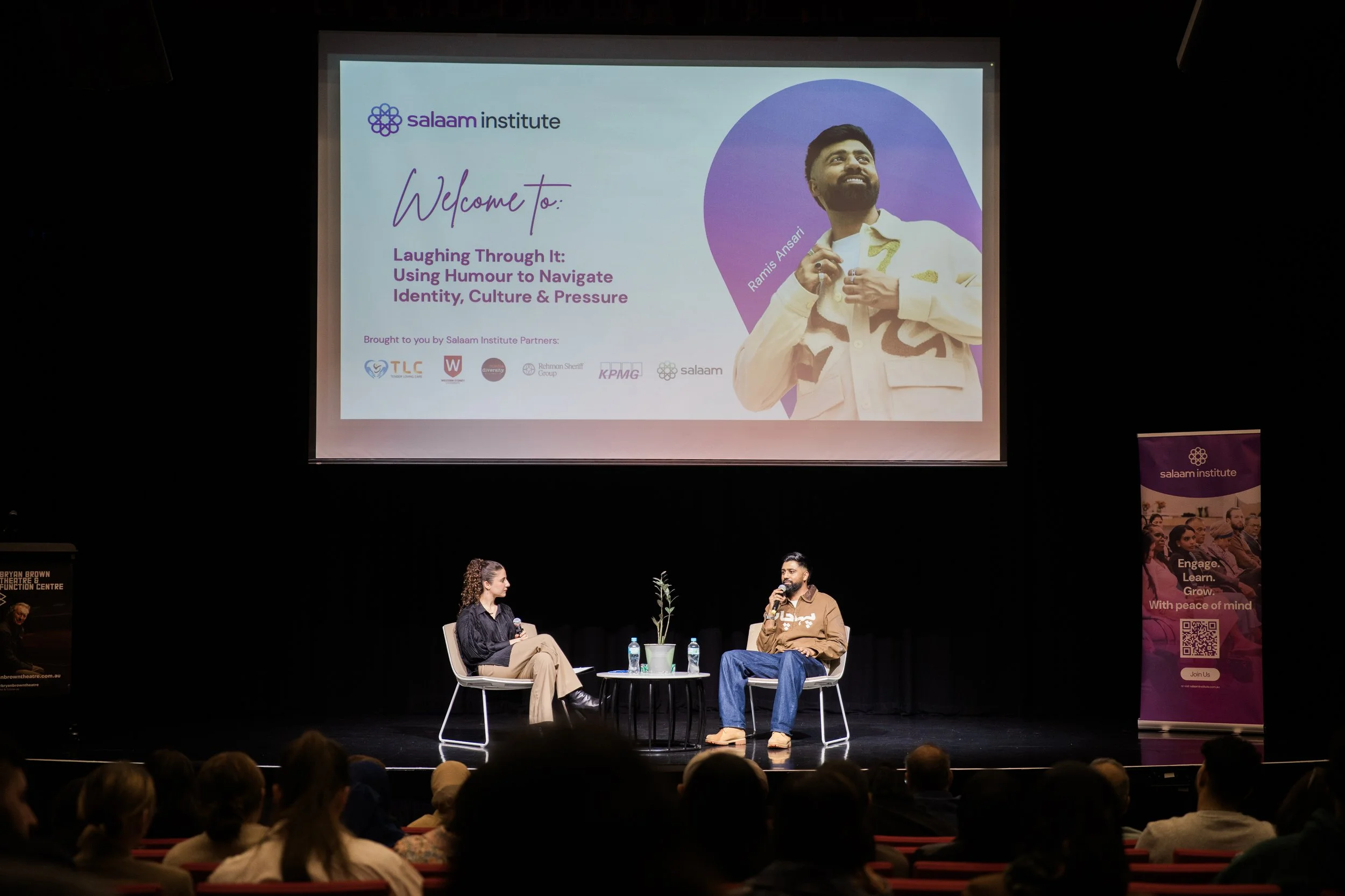 A stage with two speakers seated in chairs, one woman and one man, holding microphones while engaging in a discussion. Behind them, a large screen displays a presentation slide with the Salaam Institute logo, a photo of Ramis Ansari, and text about humor and identity. Audience members are visible in the foreground.