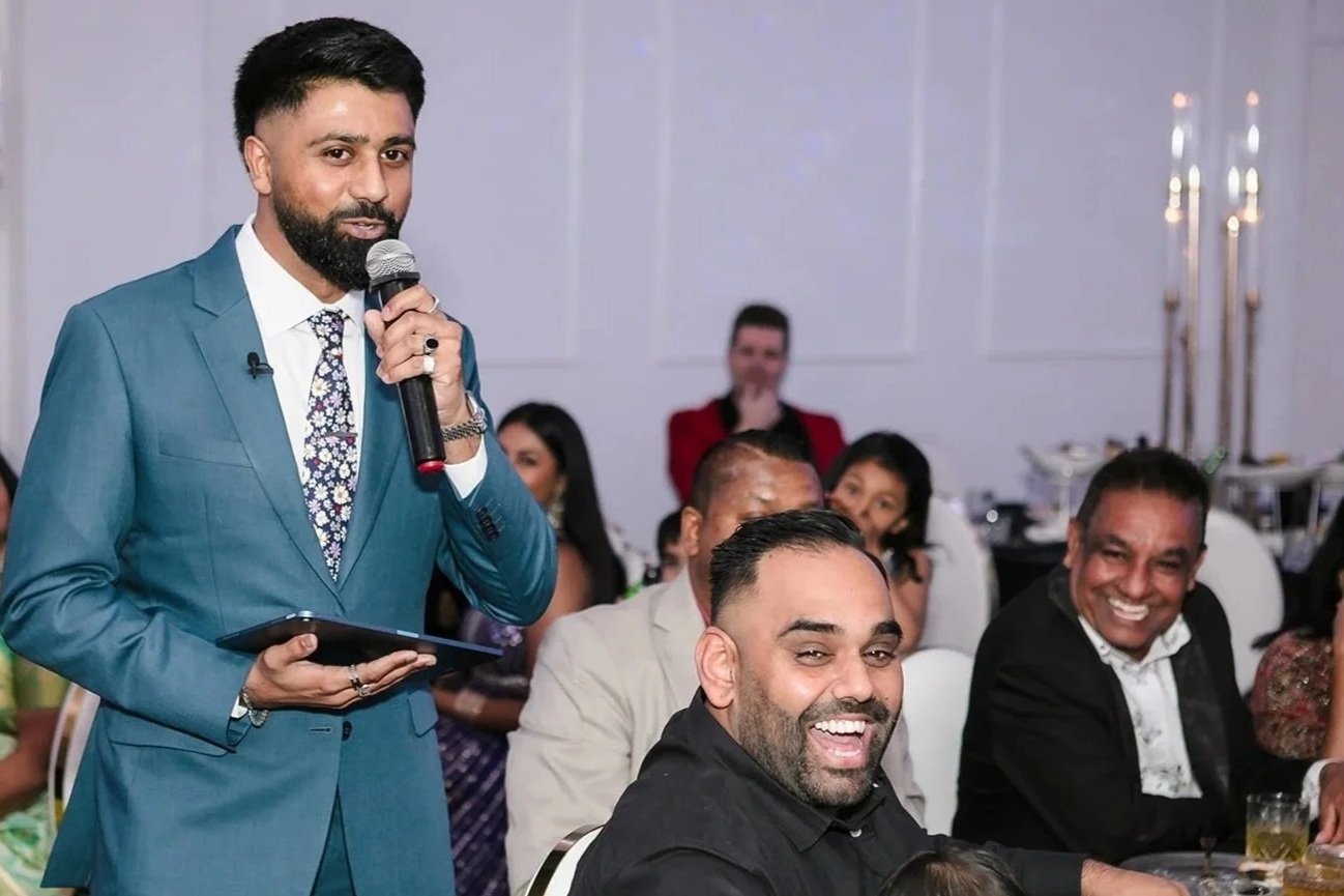 Man in a blue suit speaking into a microphone at a formal gathering surrounded by seated guests smiling and listening.