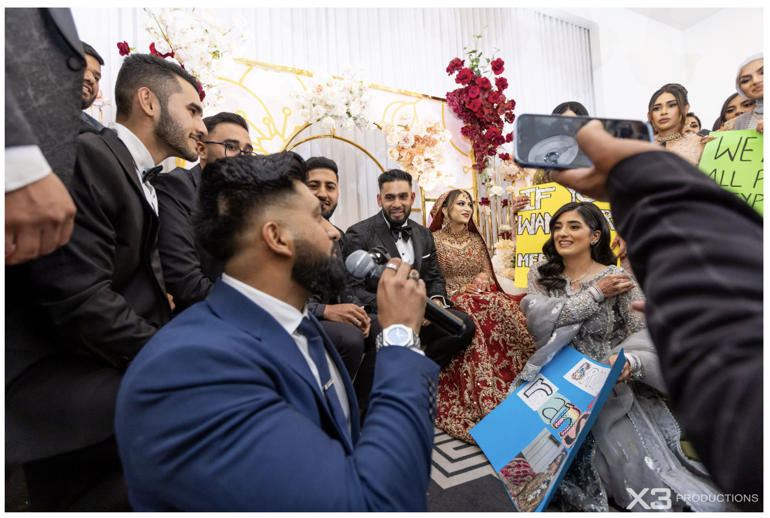 A group of people dressed in formal attire, including traditional South Asian clothing, participating in a wedding or cultural event. They are sitting and holding signs, with a decorated floral backdrop in the background.