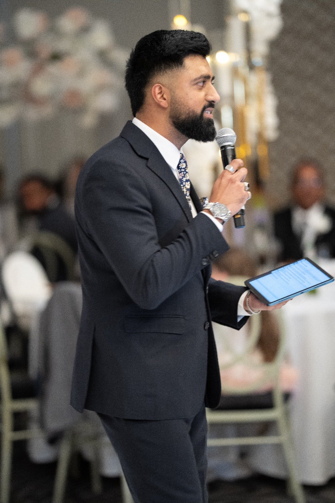 A man in a suit holding a microphone and a tablet while speaking at an event.