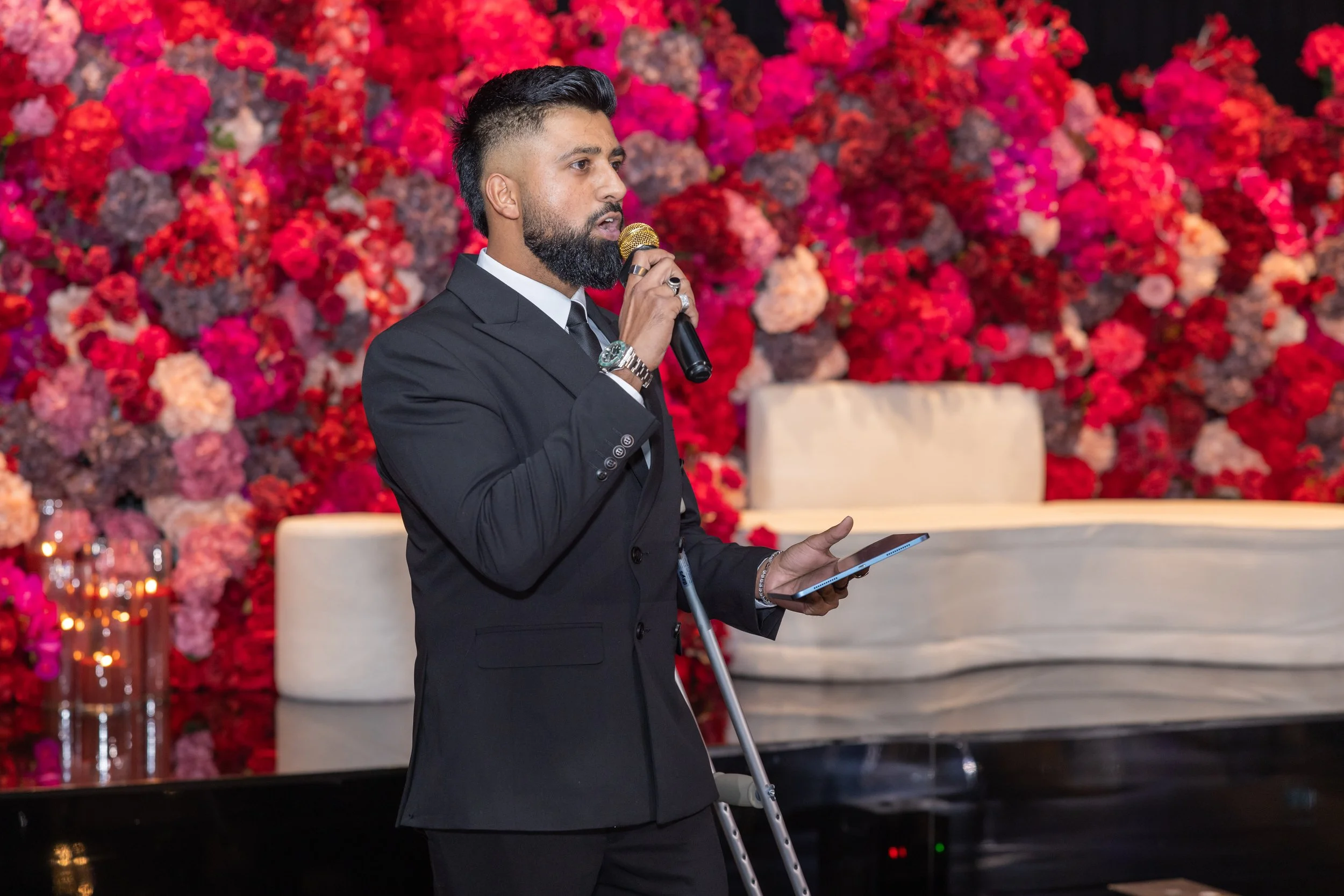 Man with beard in black suit holding a microphone and a tablet, speaking at an event with a backdrop of vibrant pink and red flowers.
