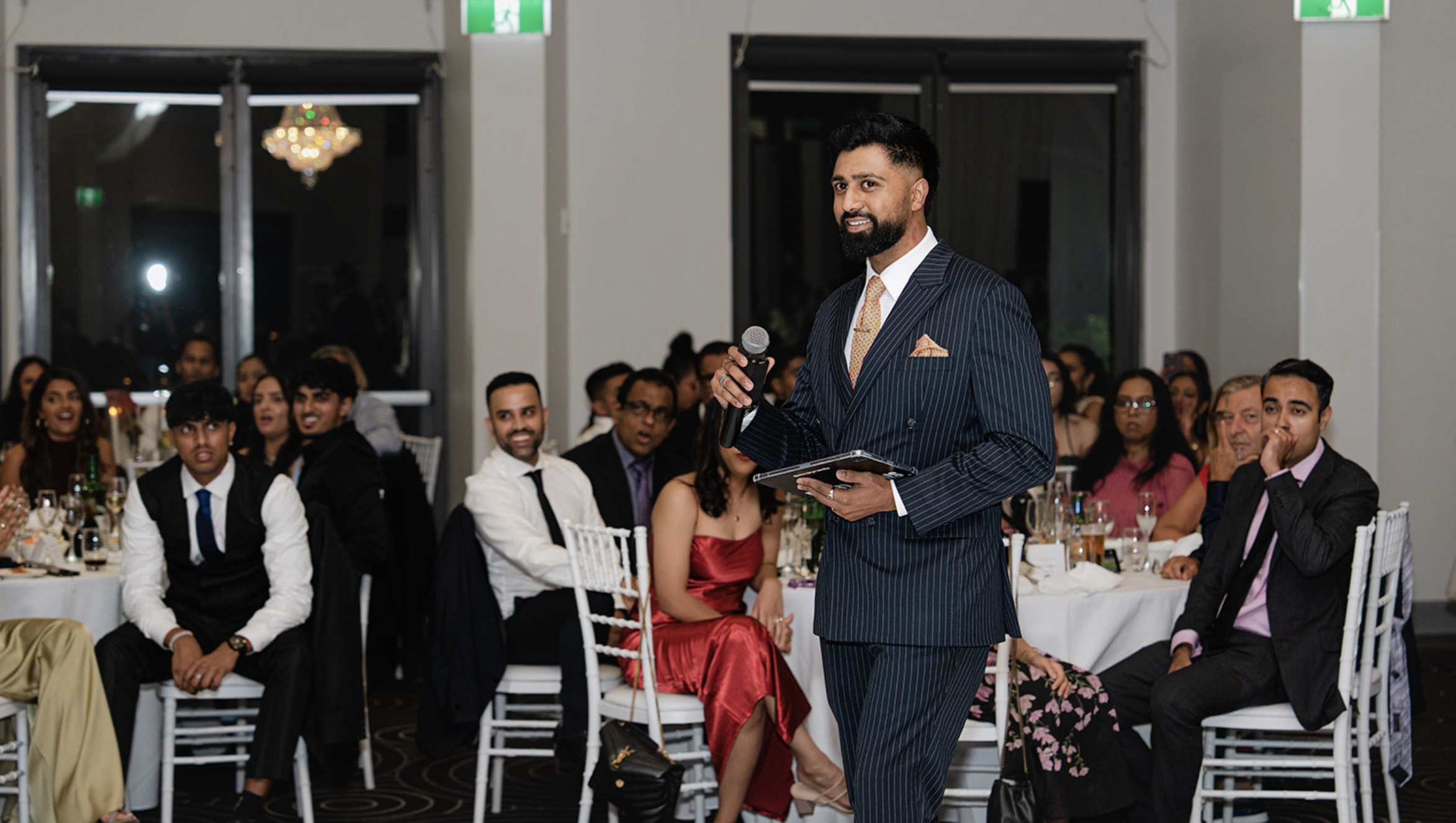 A man in a dark pinstripe suit holding a microphone and a tablet speaking at a formal event, with a diverse group of seated guests watching, some smiling and others listening, in a well-lit venue with large windows and elegant decor.