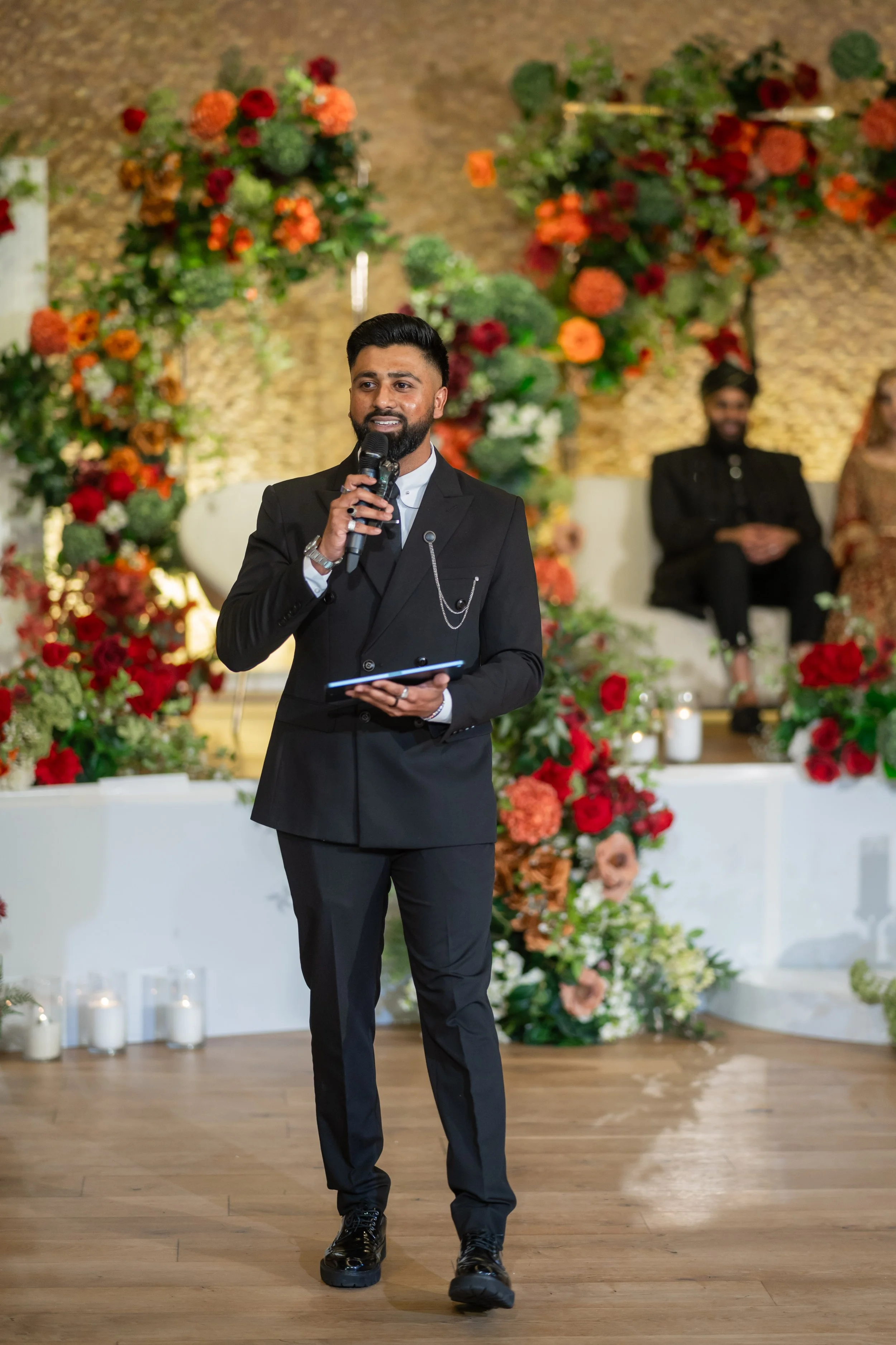 A man in a black suit holding a microphone and speaking at a wedding or formal event, with a floral backdrop and seated guests in the background.