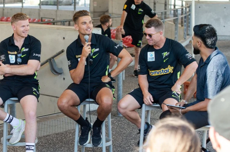 Group of sports players and a interviewer sitting in a semi-circle during a discussion at an outdoor event.